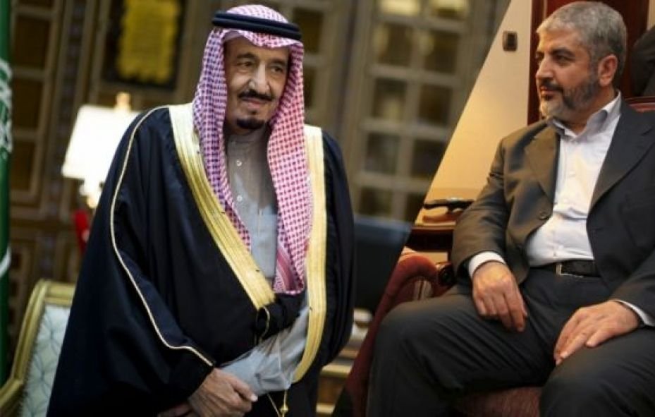 تظهر الصورة شخصين يجلسان في بيئة رسمية. الشخص على اليسار يرتدي لباساً تقليدياً يتضمن عقالاً وعباءة، مما يشير إلى هويته الثقافية. الشخص على اليمين يرتدي بدلة رسمية. يبدو أنهما يتبادلان الحديث في سياق دبلوماسي أو رسمي. الخلفية تضم تفاصيل معمارية تشير إلى مكان مهم.