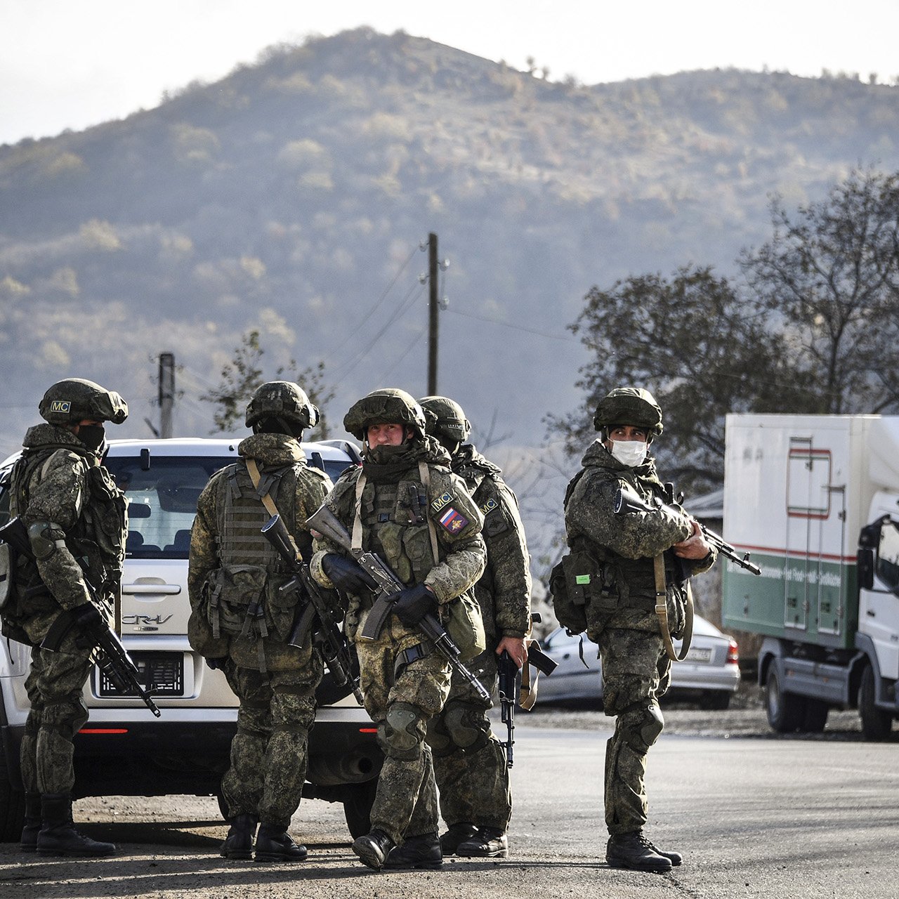 L'image montre un groupe de soldats armés en uniforme, se tenant près d'un véhicule. Ils semblent être en position de vigilance, avec des montagnes en arrière-plan. L'atmosphère est sérieuse, suggérant une opération militaire ou une situation de sécurité. Des camions peuvent également être aperçus à proximité, ajoutant un contexte logistique à la scène.