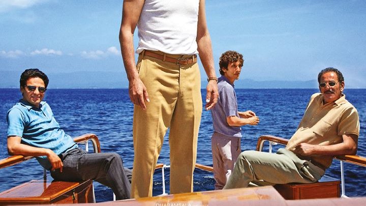 L'image montre quatre hommes sur un bateau, prenant la pose avec un fond marin. Le personnage central, portant un débardeur blanc, semble confiant et déterminé, tandis que les autres hommes sont assis autour de lui. Leurs expressions reflètent une atmosphère d'espoir et de camaraderie. Le texte mentionne "Algérie 1962, l'avenir est devant eux" et indique qu'il s'agit d'un film intitulé "L'Oranais", réalisé par Lyes Salem.