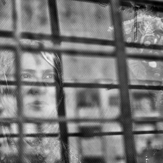 L'image montre une femme qui apparaît derrière une grille. Le contraste entre le flou et les barreaux de la grille donne une impression de séparation ou d'enfermement. La femme semble contemplative, et son expression peut évoquer des émotions profondes. Les nuances de noir et blanc ajoutent une atmosphère mélancolique à la scène.