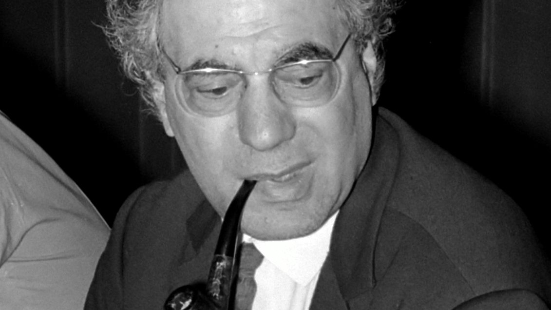 The image features a man seated at a table, wearing a suit and tie. He has a full head of curly hair and is holding a pipe in his mouth, which gives him a thoughtful or contemplative appearance. The setting seems to be a formal or professional environment, likely during a meeting or discussion. The lighting is subdued, which adds to the serious tone of the scene.