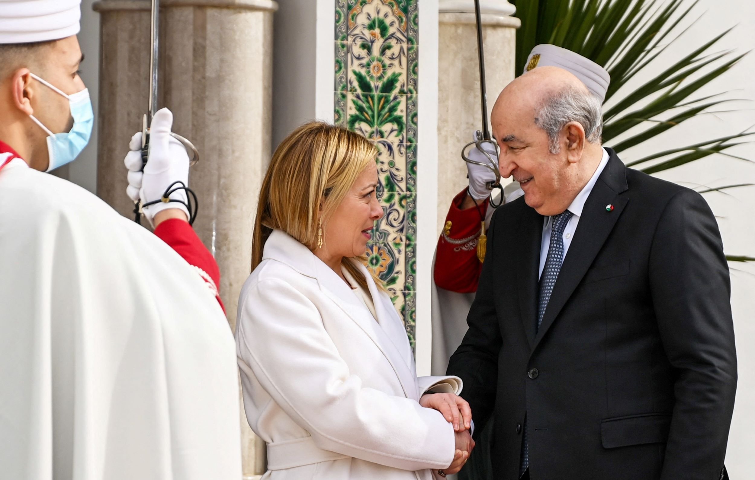 L'image montre deux personnes se serrant la main avec des sourires. Une femme, habillée en blanc, semble accueillante, tandis qu'un homme en costume sombre lui répond avec un sourire. En arrière-plan, on peut voir des gardes en uniforme et des éléments d'architecture décorative, ce qui suggère un contexte officiel, probablement une rencontre diplomatique. L'atmosphère est chaleureuse et conviviale.