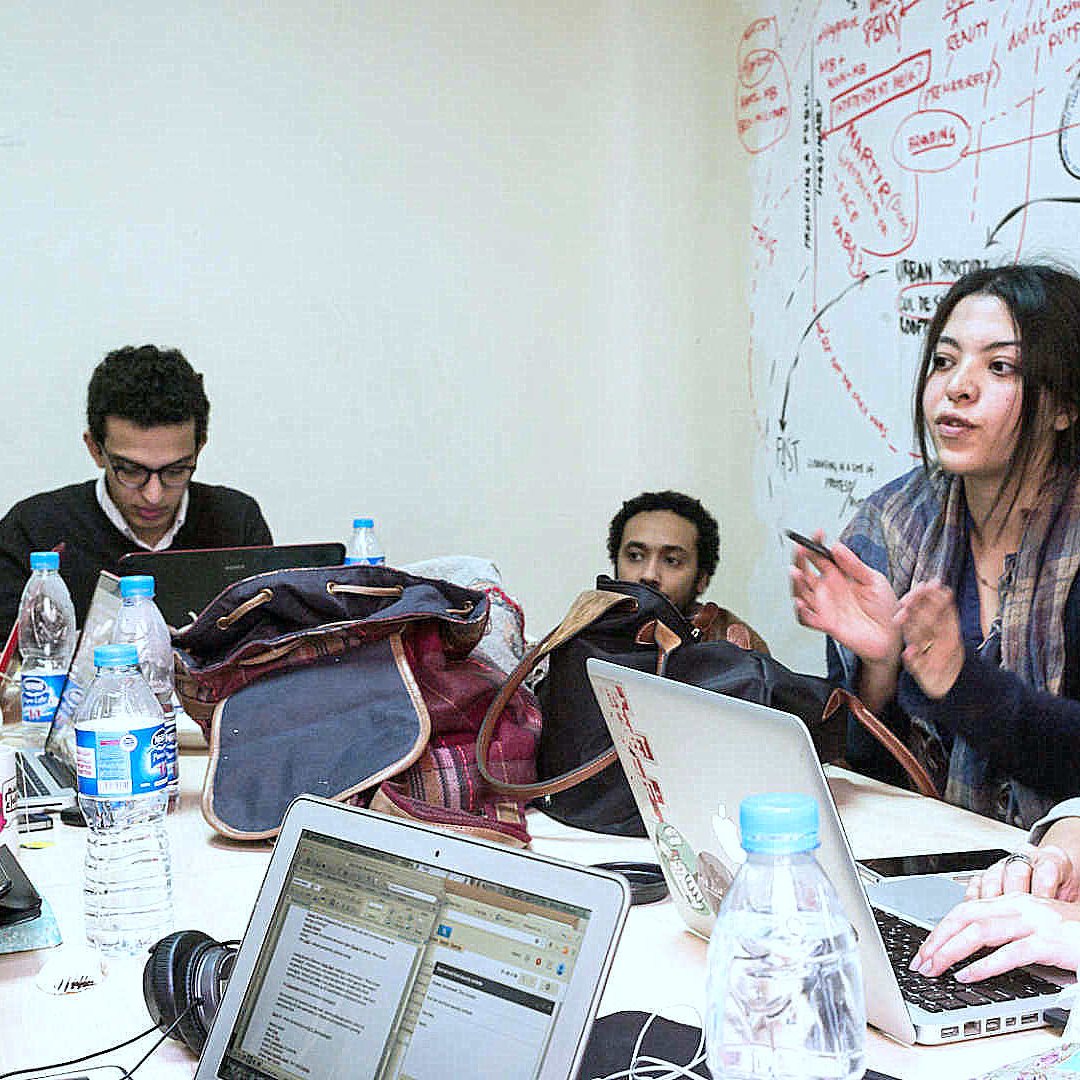 L'image montre un groupe de personnes assises autour d'une table, travaillant sur des ordinateurs portables. Il y a des bouteilles d'eau et des sacs dispersés sur la table. Une femme semble expliquer quelque chose avec enthousiasme, tandis que les autres écoutent attentivement. Le mur derrière eux est couvert de notes et de schémas, suggérant une ambiance créative et collaborative.