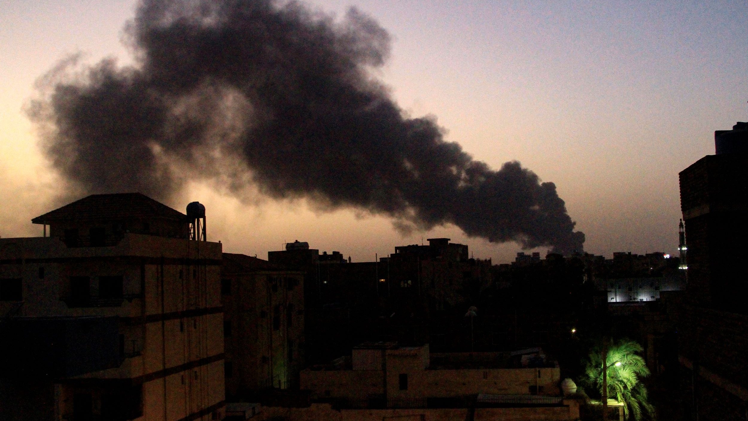 L'image montre un paysage urbain de nuit, avec des bâtiments au premier plan. En arrière-plan, de grands nuages de fumée noire s'élèvent dans le ciel, créant un contraste avec l'horizon teinté de lumière douce. On peut observer une ambiance sombre et impressionnante, suggérant une situation potentiellement problématique ou alarmante. Les silhouettes des bâtiments et les arbres ajoutent de la profondeur à la scène.