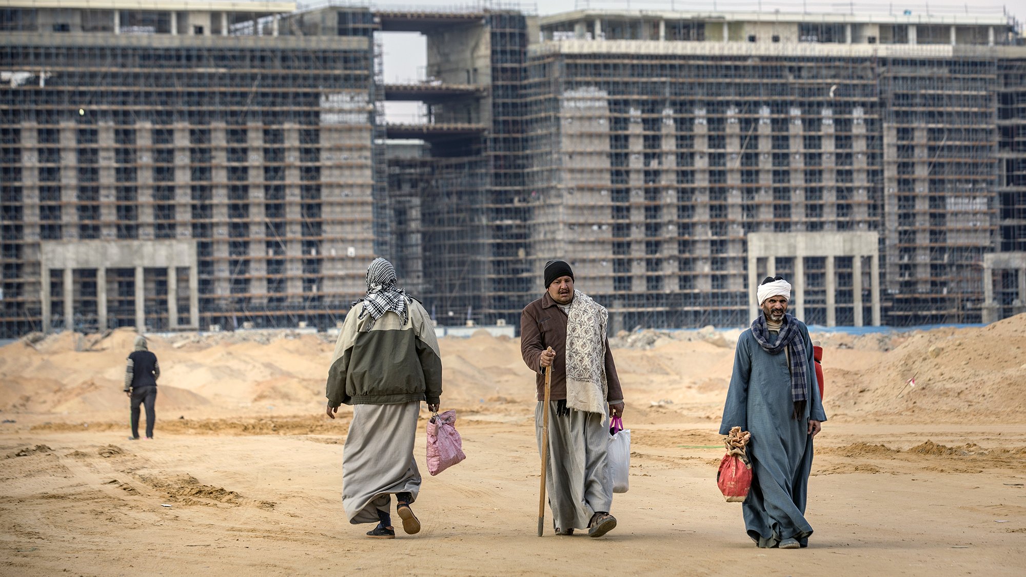 L'image montre un paysage de construction, avec plusieurs bâtiments inachevés en arrière-plan. Au premier plan, on voit quatre personnes marchant sur un sol de terre. Elles portent des vêtements traditionnels et semblent transporter des sacs. L'environnement est désertique, avec des matériaux de construction visibles autour. Le ciel est nuageux, contribuant à l'atmosphère de travail en cours.