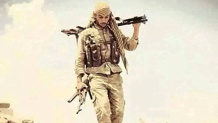 L'image montre un homme portant une tenue militaire, avec un chèche sur la tête. Il marche sur un terrain désertique, tenant deux armes, l'une à la main et l'autre portée sur son épaule. Son expression semble déterminée et il est entouré d'un paysage aride. L'atmosphère de l'image évoque un contexte de conflit ou de guerre.