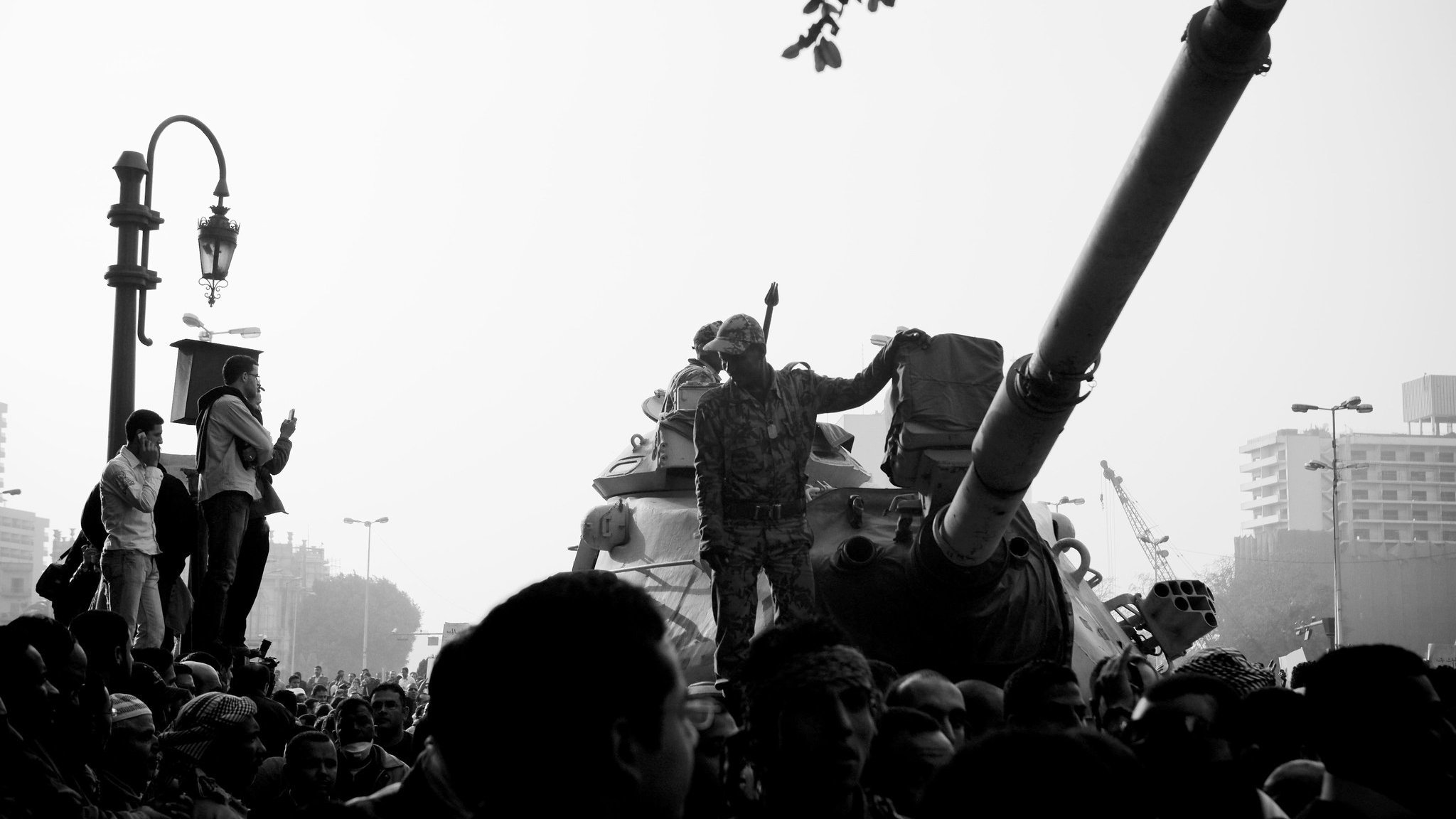 L'image montre une scène de manifestation ou de rassemblement avec une atmosphère intense. Au centre, on peut voir un soldat debout sur un char, levant le bras en signe de défi ou de puissance. Le char est entouré d'une foule dense de personnes, certaines levant les bras ou prenant des photos. L'arrière-plan semble urbain, avec des bâtiments et un lampadaire. La photo est en noir et blanc, ce qui ajoute à la dramaturgie et à la tension de la scène.