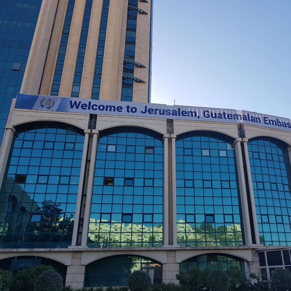 L'image montre un bâtiment moderne, probablement un immeuble administratif, avec une façade en verre réfléchissant. Au-dessus des fenêtres, une grande banderole affiche le message "Welcome to Jerusalem, Guatemalan Embassy". Le ciel est dégagé, suggérant une belle journée. Des arbres et des plantes ornent le devant du bâtiment, ajoutant une touche de verdure à l'environnement urbain.
