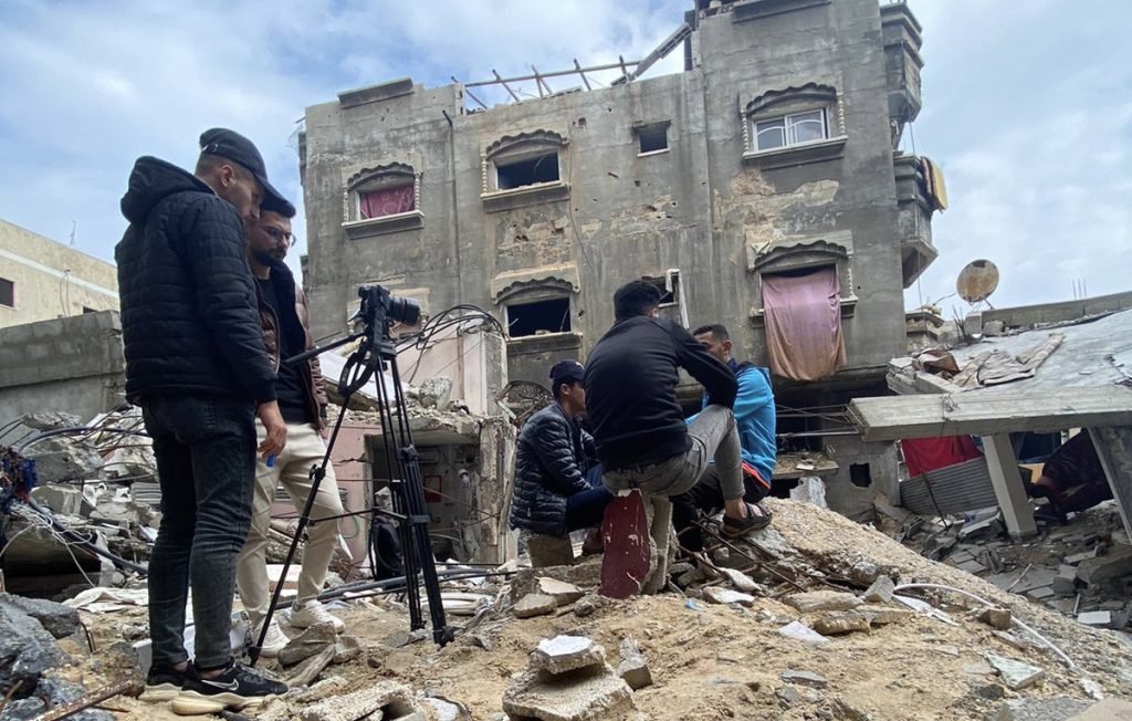 L'image montre un groupe de personnes se tenant sur des décombres, probablement après un effondrement de bâtiment. Ils semblent être en train de filmer ou de prendre des photos, avec du matériel de caméra visible. En arrière-plan, on peut voir une structure endommagée, avec des fenêtres brisées et des pans de mur manquants. Le ciel est nuageux, ce qui ajoute à l'atmosphère sombre de la scène. L'environnement est chaotique, reflétant les conséquences d'une destruction.