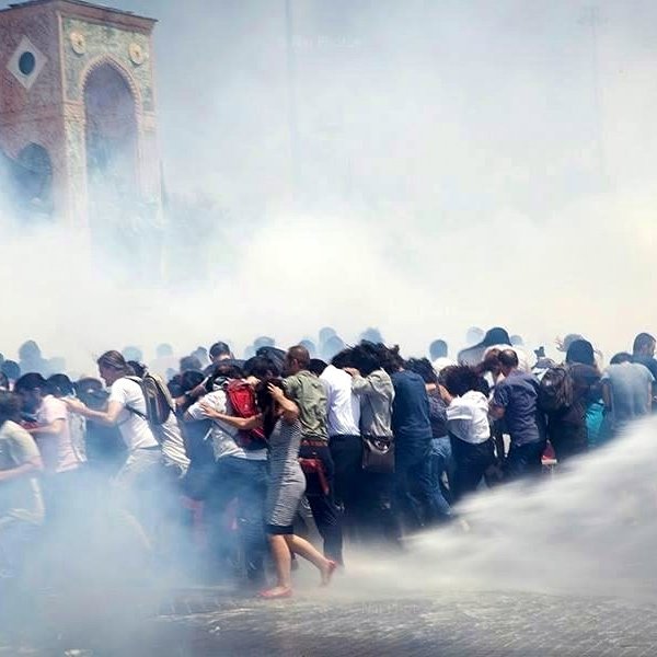 L'image montre une scène de manifestation où un grand groupe de personnes semble être en mouvement, probablement en train d'échapper à des gaz lacrymogènes ou à une intervention des forces de l'ordre. Le brouillard blanc, causé par la fumée ou les sprays, rend l'atmosphère tendue et chaotique. En arrière-plan, une structure architecturale est visible, mais elle est à peine discernable à cause de la fumée. Les manifestants semblent rassemblés et stressés, certains essayant de se protéger ou de se diriger vers une zone plus sûre.