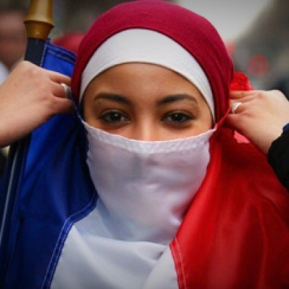 The image features a young woman dressed in a red and white outfit, with a hijab, who is adjusting her clothing. She is holding a flag that represents France, with the colors of blue, white, and red. The background suggests a public gathering or demonstration, with people present in a city setting. The atmosphere appears to convey themes of identity and cultural expression.