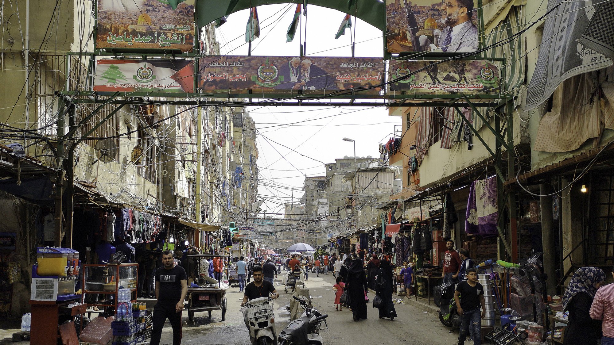 L'image montre une rue animée d'un quartier urbain. On peut y voir des bâtiments étroits et des fils électriques visibles. Des drapeaux et des affiches, probablement liés à un événement ou à une célébration, décorent l'entrée de la rue. Des personnes circulent, certaines à pied et d'autres à moto, ajoutant à l'activité de la scène. Le lieu semble être un marché ou un centre commercial, avec des étals vendant divers produits. L'atmosphère est vibrante et vivante.