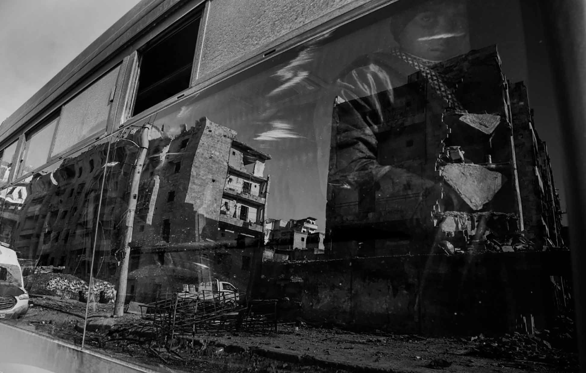 الصورة تظهر منظرًا حضريًا بواسطة زجاج نافذة حافلة. تعكس الصورة مجموعة من المباني المتضررة، مما يدل على دمار أو تدهور في المنطقة. يظهر أيضاً شخص يجلس بجانب النافذة، وملامح وجهه غير واضحة بسبب الانعكاس. الألوان في الصورة تتسم باللونين الأبيض والأسود، مما يضفي طابعاً درامياً وأجواءً حزينة على المشهد.