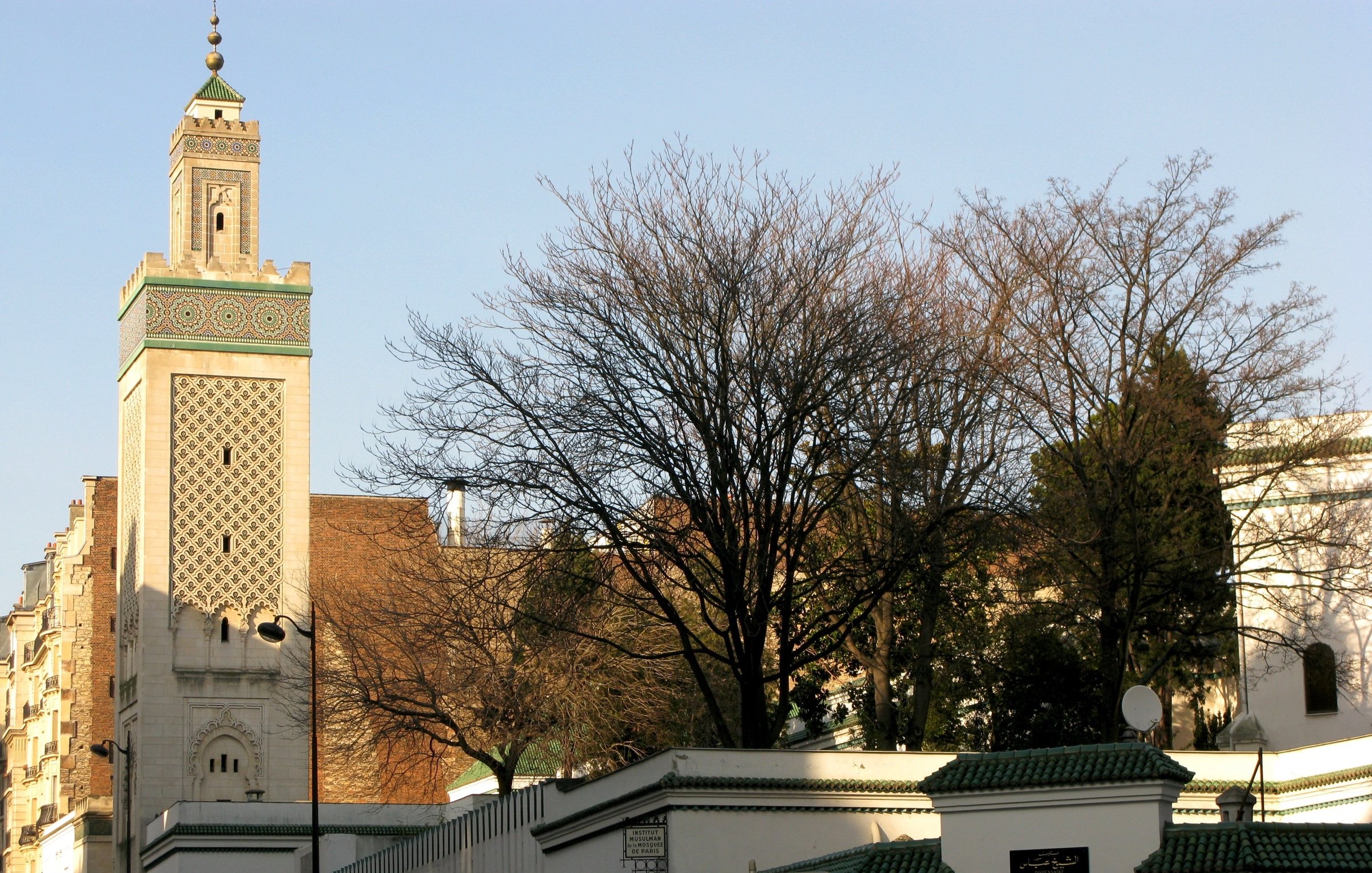 L'image montre une vue d'un bâtiment architectural distinctif, probablement une mosquée, avec un minaret élancé décoré de motifs. En arrière-plan, on aperçoit des arbres dépouillés, ce qui indique peut-être une saison hivernale ou automnale. Le mur entourant le bâtiment est blanc avec des bordures vertes. Des personnes marchent le long de la rue, créant une atmosphère de vie urbaine. Le ciel est dégagé, suggérant une journée ensoleillée.