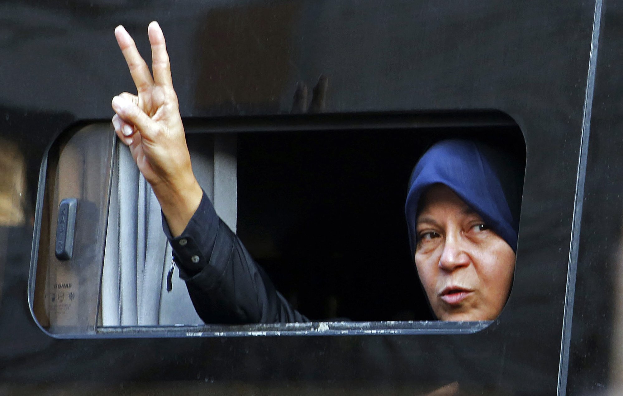 L'image montre une femme qui fait un signe de paix avec sa main, en levant deux doigts. Elle est visible à travers une fenêtre légèrement ouverte d'un véhicule. La femme porte un hijab. Son expression semble déterminée et engagée. Le fond est sombre, ce qui met en avant son geste et son visage.