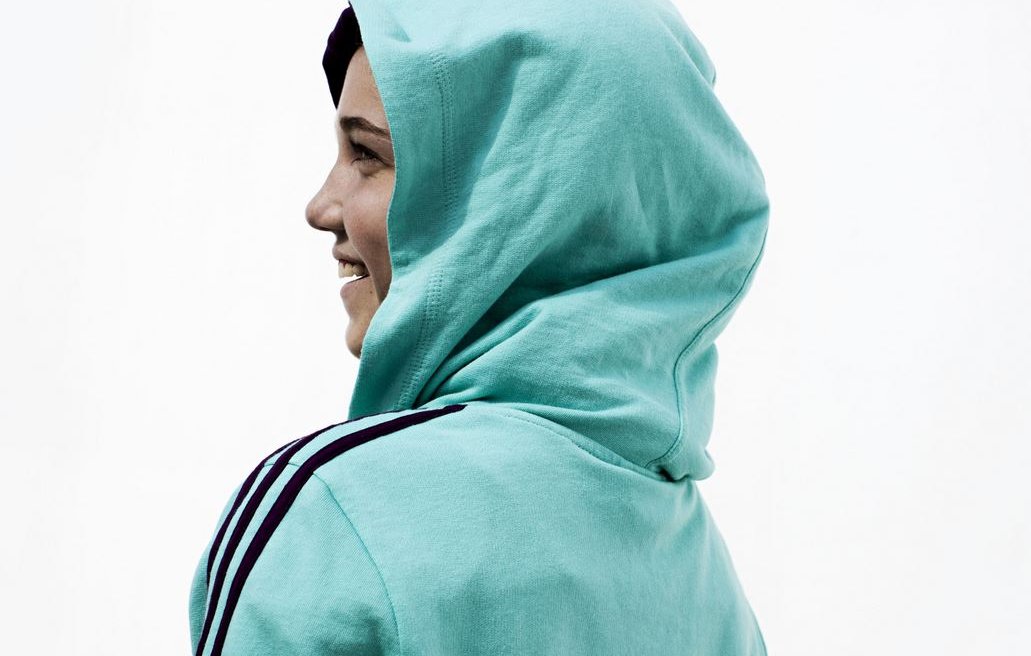 L'image montre une personne de profil, portant un sweat à capuche de couleur turquoise. Le sweat arbore le mot "QATAR" inscrit à l'arrière. La personne semble sourire, et la capuche est relevée sur sa tête. L'arrière-plan est neutre, mettant l'accent sur la tenue de la personne.
