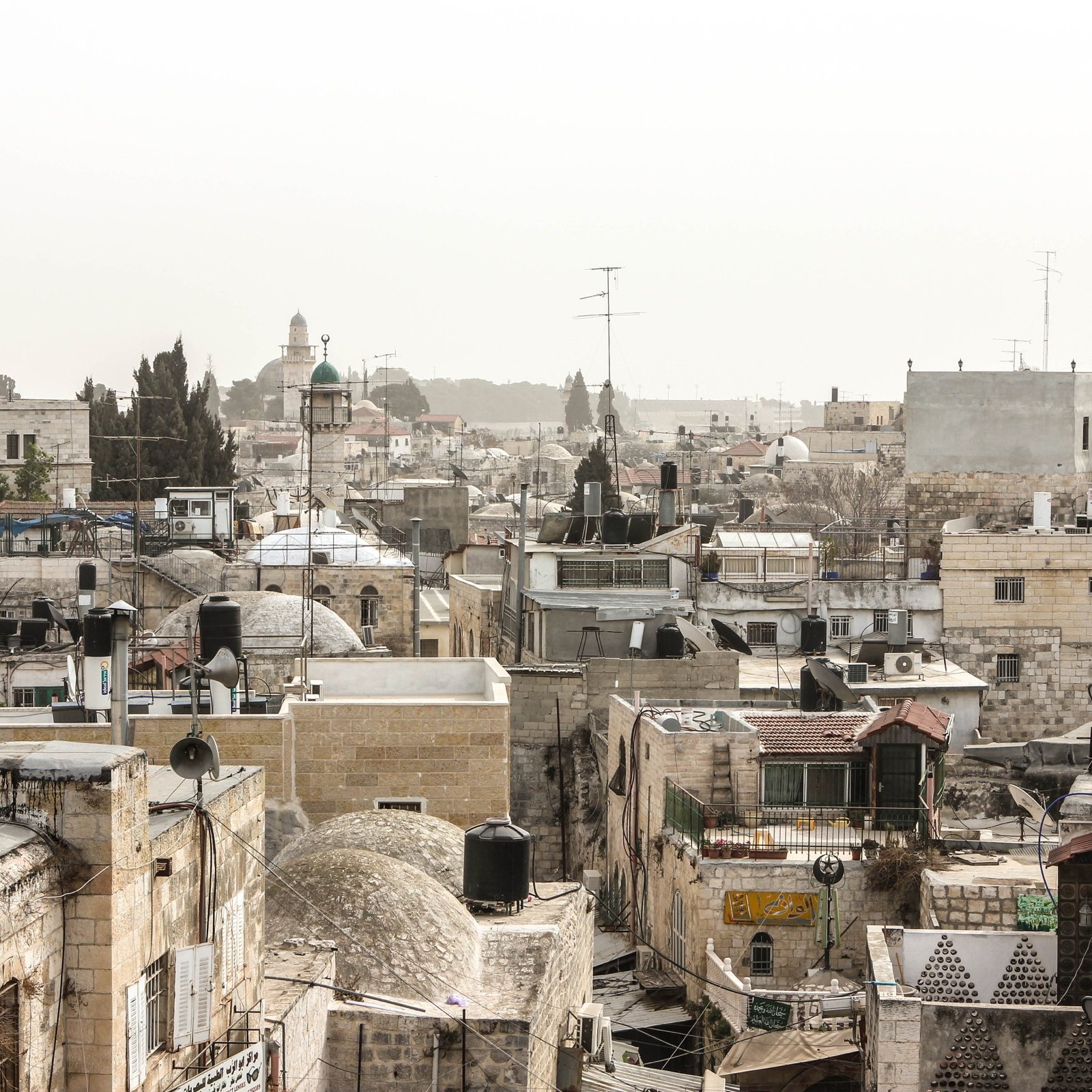 الصورة تُظهر منظرًا لأحد الأحياء القديمة، حيث تتداخل المباني التقليدية مع الأسطح المسطحة وأجهزة التكييف. يوجد أيضًا بعض الأشجار ومآذن المساجد في الخلفية، مما يشير إلى طابع تاريخي وديني للمنطقة. السماء تبدو مائلة إلى الرمادي، مما قد يدل على أجواء غائمة أو ضبابية.