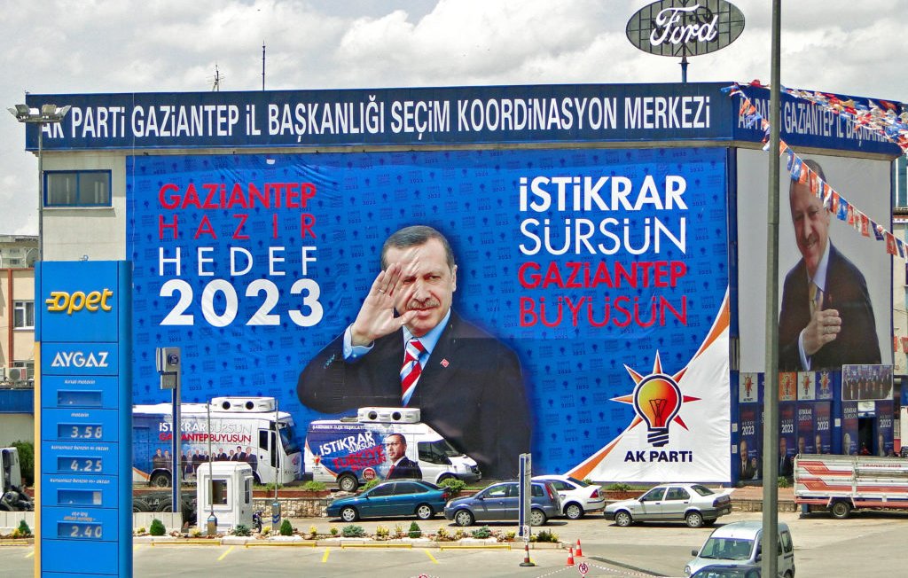 L'image montre un grand panneau publicitaire avec des slogans politiques. On y voit des phrases comme "GAZİANTEP HAZIR 2023" et "İSTİKRAR SÜRSÜN GAZİANTEP BÜYÜSÜN", suggérant une campagne électorale pour les élections de 2023. L'image présente également une figure politique qui salue, ajoutant une dimension dynamique à la publicité. Le fond inclut des symboles ou logos associés à un parti, probablement le parti AK. La scène est typique des campagnes électorales visuelles, avec une mise en avant forte du message et de la personnalité politique.