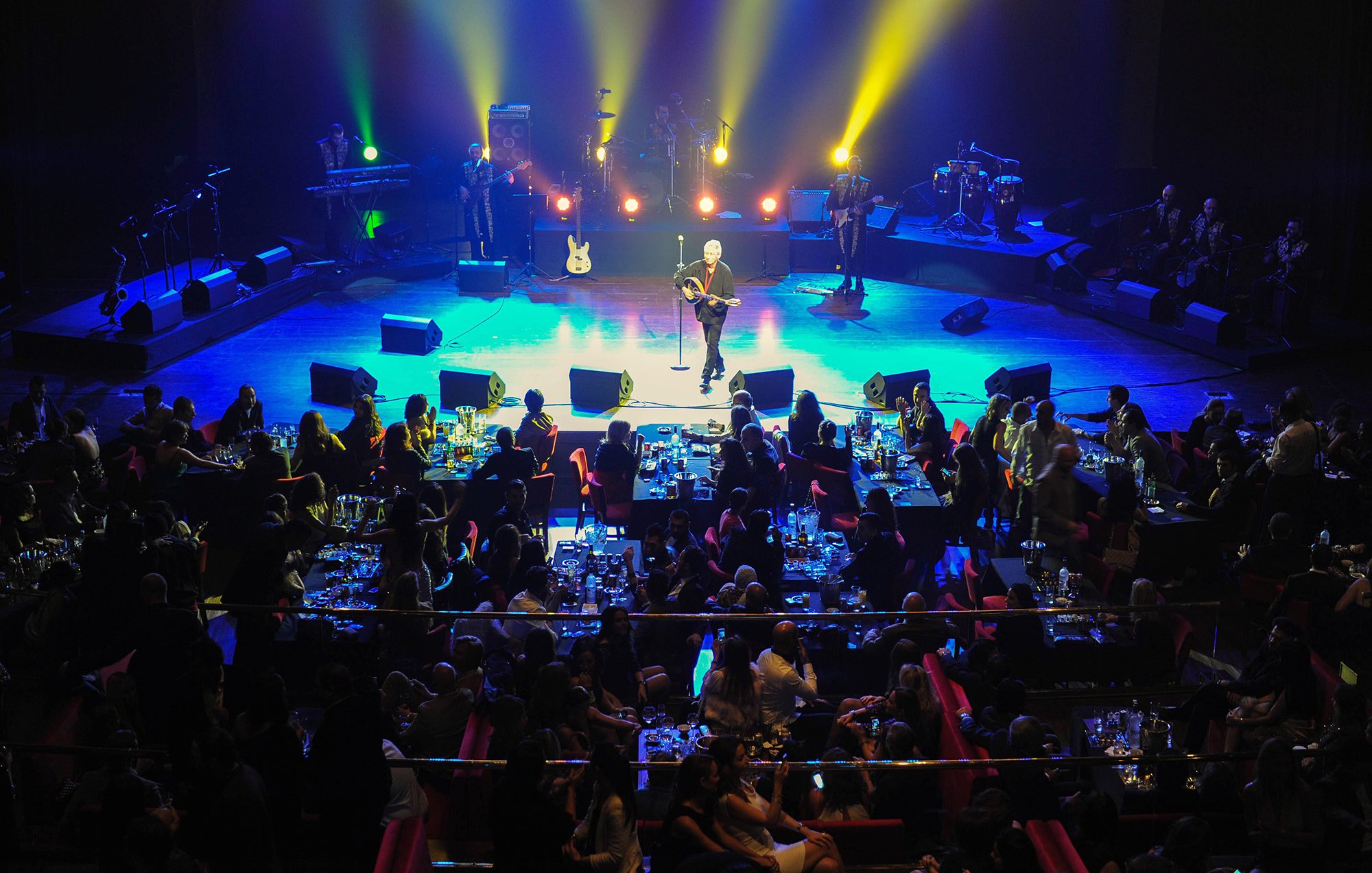 L'image montre une scène de concert animée. Au premier plan, un artiste se produit sur scène, entouré de lumières colorées et de spots éclairant l'espace. En arrière-plan, on peut voir des musiciens qui l'accompagnent. La salle est pleine de spectateurs assis à des tables, savourant leur soirée avec des repas et des boissons. L'ambiance est festive et chaleureuse, avec des gens qui semblent apprécier la performance.