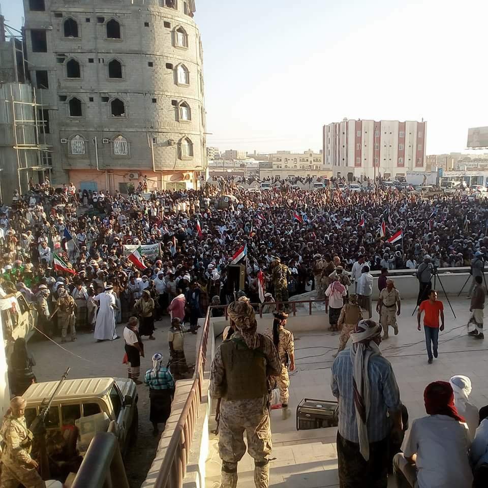 L'image montre une grande foule rassemblée lors d'une manifestation ou d'un rassemblement public. Les participants tiennent des banderoles et des drapeaux, indiquant un fort engagement politique ou social. En arrière-plan, on peut voir des bâtiments en construction ou rénovés, ainsi qu'une architecture variée. La scène semble se dérouler en plein air, probablement durant l'après-midi ou le début de la soirée, avec une lumière naturelle éclatante. L'atmosphère est dynamique, avec beaucoup de gens engagés dans une cause commune.