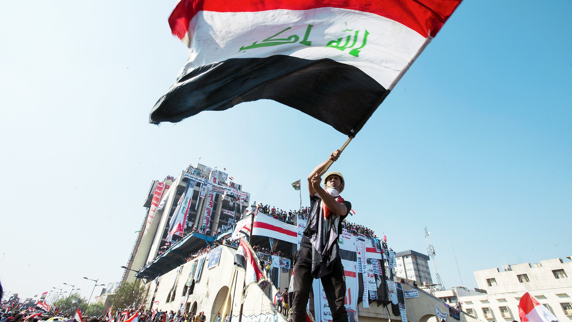 L'image montre une personne agitant un drapeau irakien. Le drapeau, aux couleurs rouge, blanc, noir et vert, est bien visible. La scène se déroule probablement lors d'une manifestation ou d'un événement public, avec des bâtiments en arrière-plan décorés de drapeaux. L'atmosphère semble dynamique, reflétant un sentiment de patriotisme et de rassemblement.