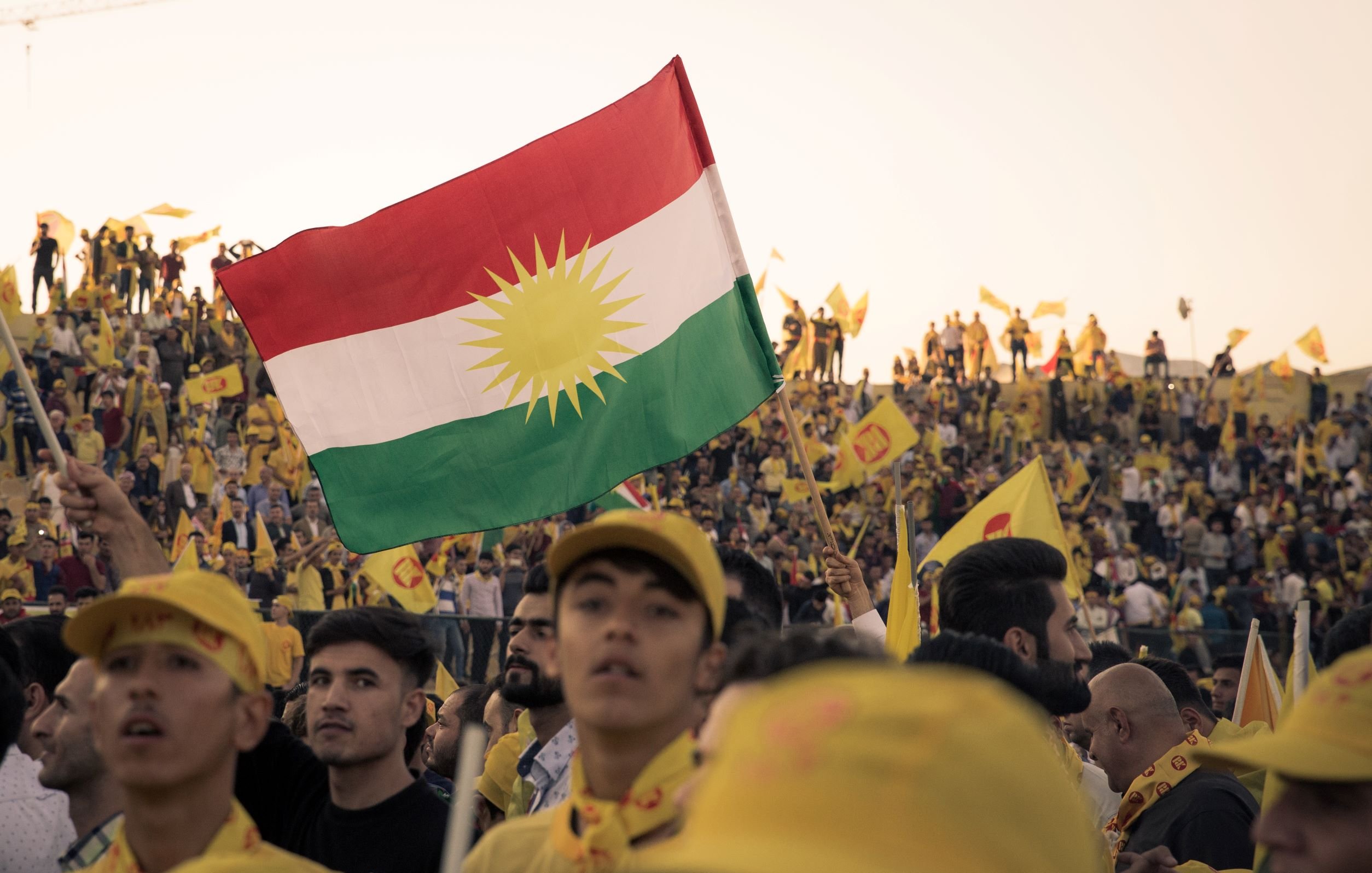 در این تصویر جمعیتی بزرگ در حال جشن و تظاهرات دیده می‌شود. تعدادی از افراد پرچم کردستان را در دستان خود دارند که شامل رنگ‌های سبز، سفید و قرمز است و در وسط آن خورشیدی قرار دارد. همچنین، جمعی از افراد کلاه‌ها و لباس‌های زرد رنگ به تن دارند و پرچم‌هایی با رنگ زرد و نمادهایی دیگر در دست دارند. فضای کلی تصویر پر از اشتیاق و انرژی مثبت است و نشان‌دهنده حمایت از یک cause یا هویت جمعی است.