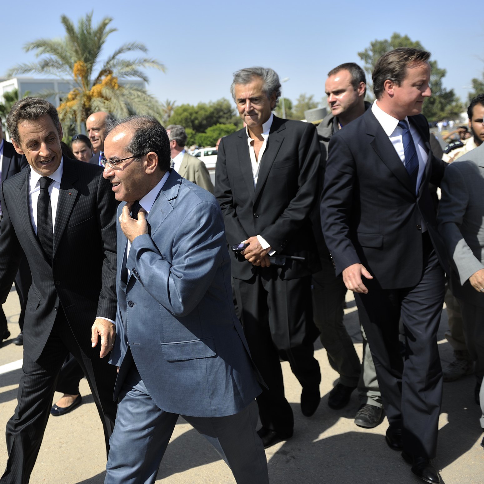 L'image montre un groupe d'hommes marchant ensemble dans un environnement extérieur. Ils semblent être en discussion animée. En arrière-plan, on peut apercevoir des palmiers et un bâtiment, suggérant un climat ensoleillé. Les hommes portent des costumes, ce qui indique un contexte formel ou professionnel. L'ambiance semble dynamique et engageante.