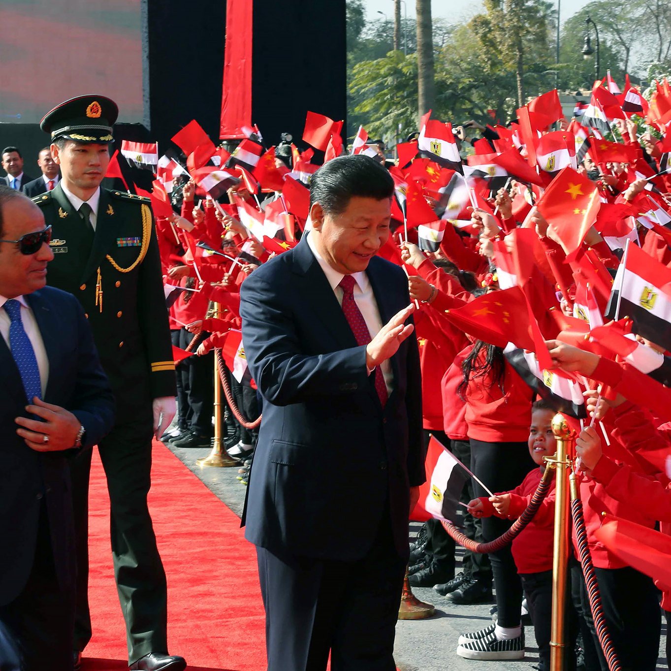 L'image montre un événement public avec des drapeaux de la Chine et de l'Égypte. Au premier plan, deux figures politiques semblent interagir avec la foule. L'un des hommes porte un costume sombre et des lunettes de soleil, tandis que l'autre est habillé d'un costume clair et sourit en saluant les personnes présentes. La foule, vêtue de vêtements rouges, tient des drapeaux et semble enthousiaste. On peut voir un tapis rouge et des gardes en uniforme à l'arrière-plan. L'ambiance est festive et accueillante.