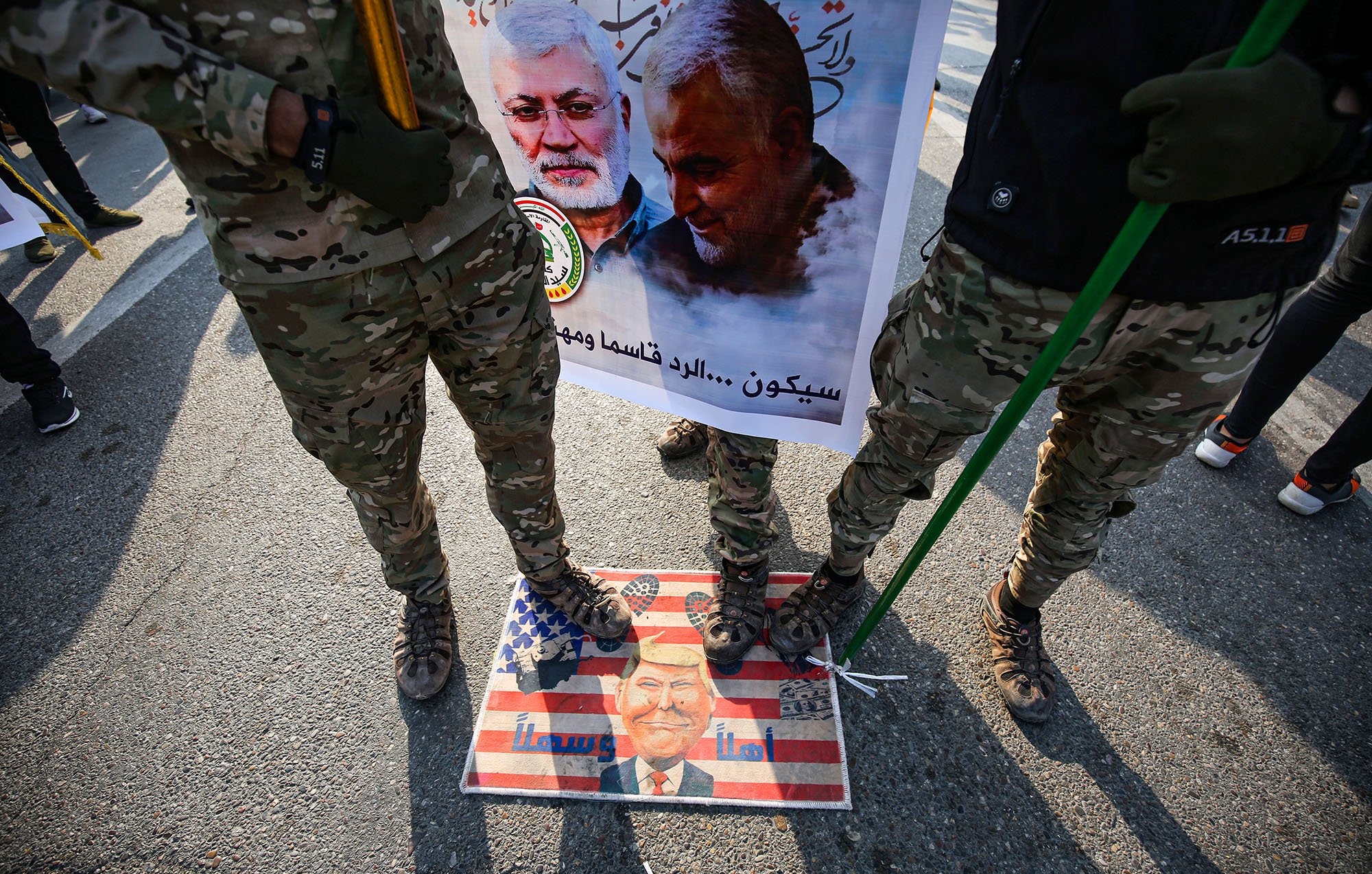 L'image montre des soldats en uniforme militaire debout sur une affiche ou un symbole qui semble représenter des sentiments anti-américains. Ils tiennent des drapeaux ou des bannières, et il y a une image de personnes, probablement des figures politiques ou militaires, sur l'affiche. Le sol est décoré d'une illustration de style artistique, mélangé avec des éléments du drapeau américain, ce qui suggère une forte opinion politique et un contexte de protestation ou de commémoration.