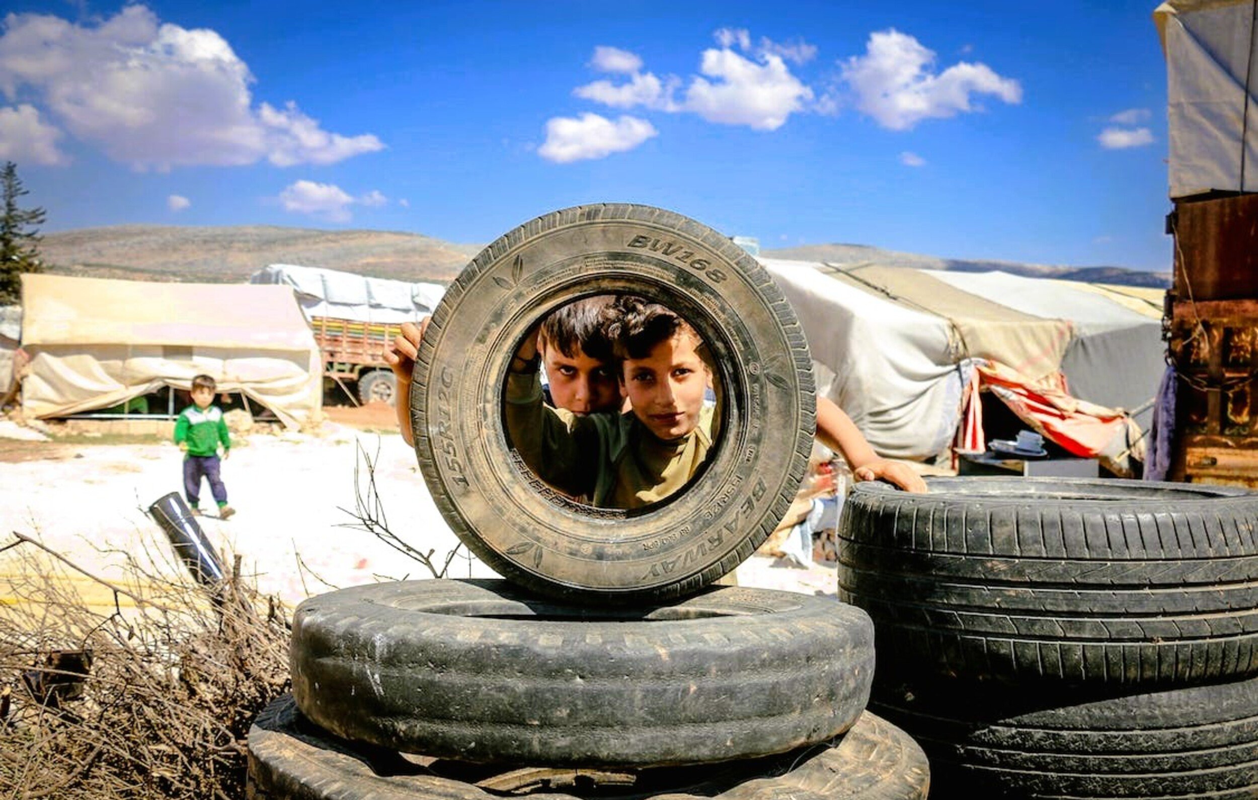 L'image montre deux enfants regardant à travers un pneu, dans un environnement qui semble être un camp de réfugiés. On peut voir des pneus empilés autour d'eux et des abris simples en arrière-plan. Le ciel est dégagé avec quelques nuages. Les enfants affichent une expression curieuse, et la scène dépeint un moment de jeu au milieu d'un cadre difficile.