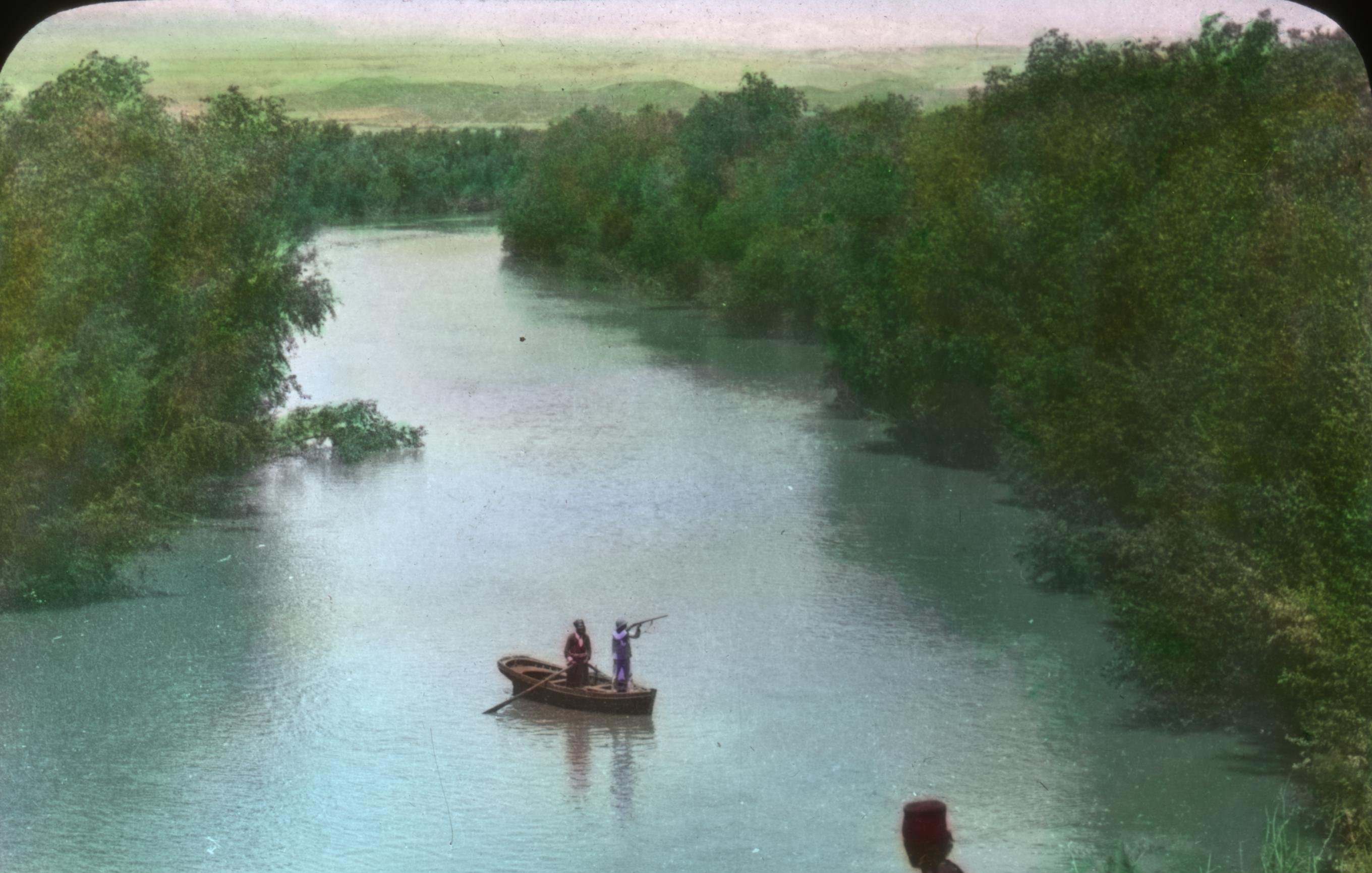 L'image montre une scène paisible au bord d'une rivière. Deux personnes se tiennent sur la berge, regardant une barque qui navigue sur l'eau. La barque semble être occupée par deux personnes qui sont en train de pêcher ou d'observer les environs. La végétation entourant la rivière est dense et verdoyante, ajoutant une touche de nature à la scène. Le ciel est clair et dégage une atmosphère calme et sereine.