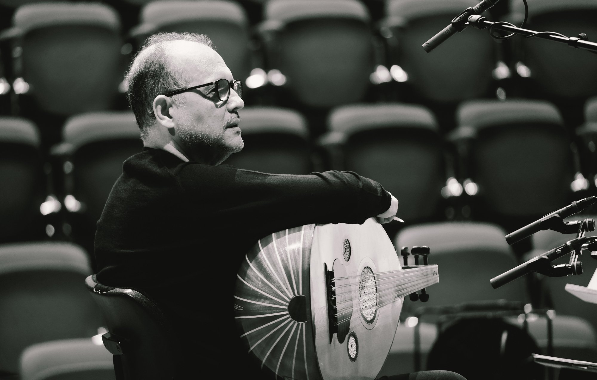 L'image montre un homme assis, tourné légèrement sur le côté. Il porte des lunettes et un pull noir. Dans ses bras, il tient un instrument de musique en forme de luth. L'arrière-plan est flou avec des rangées de sièges.