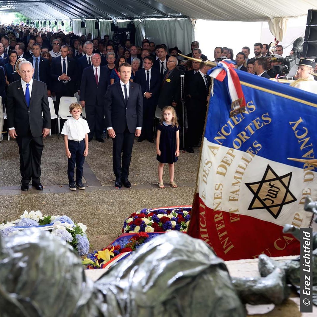 The image depicts a solemn gathering under a tent, likely at a memorial or commemorative event. In the foreground, there are several children and adults standing attentively. Prominent figures, possibly including political leaders, are present, and flags, including one with a star emblem, are displayed around them. The atmosphere appears respectful and reflective, suggesting it is a tribute to those who have passed or a celebration of their memory. Floral arrangements are visible, adding to the somber yet respectful tone of the scene.