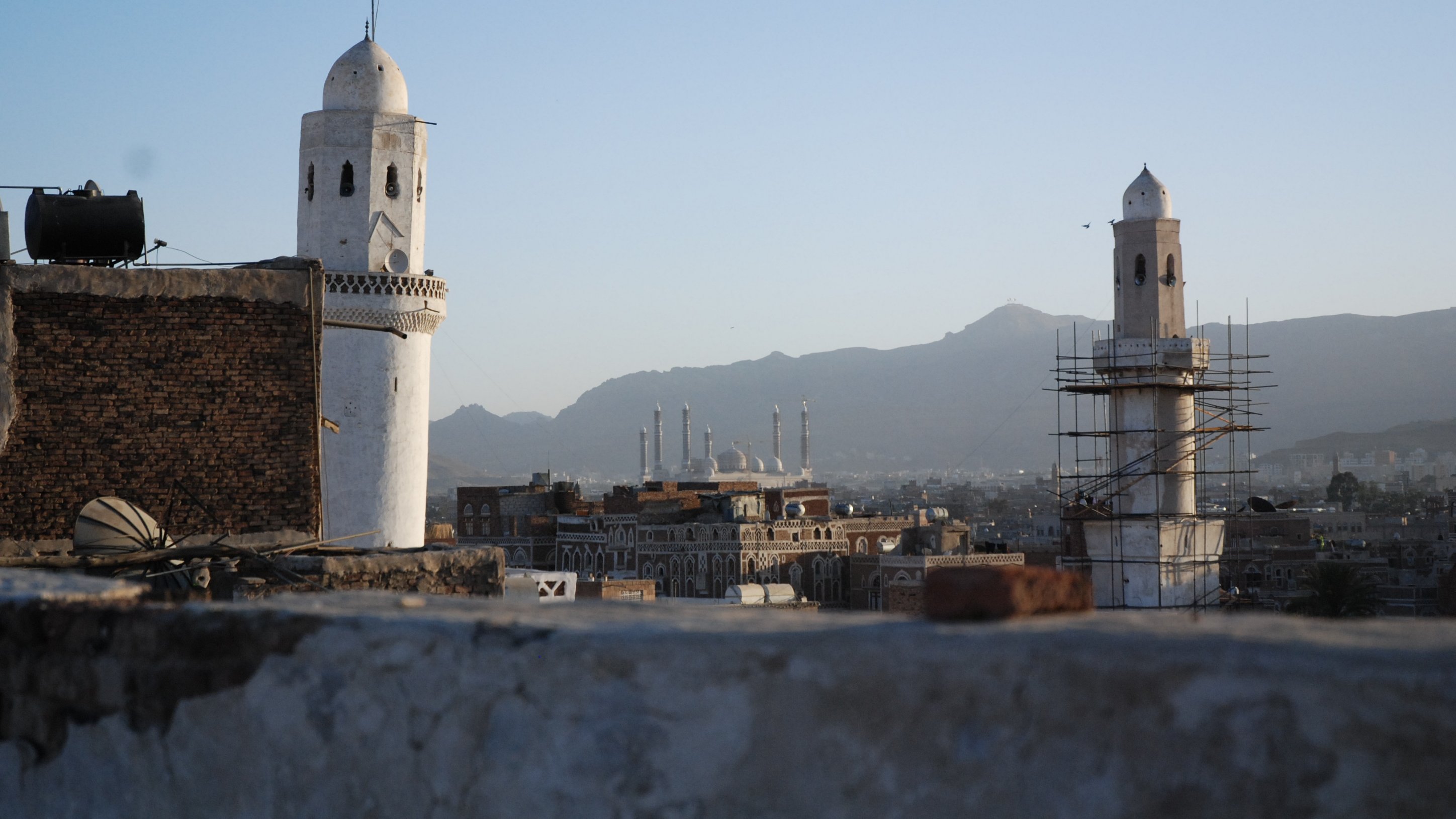 L'image montre une vue panoramique d'une ville avec des minarets distinctifs. On peut voir deux tours blanches caractéristiques, l'une étant en cours de restauration, entourées d'un paysage montagneux en arrière-plan. Le ciel est clair, indiquant une ambiance paisible au crépuscule. Au loin, plusieurs autres structures architecturales, probablement des mosquées, se dessinent à l'horizon, contribuant à l'atmosphère culturelle et historique de la scène.