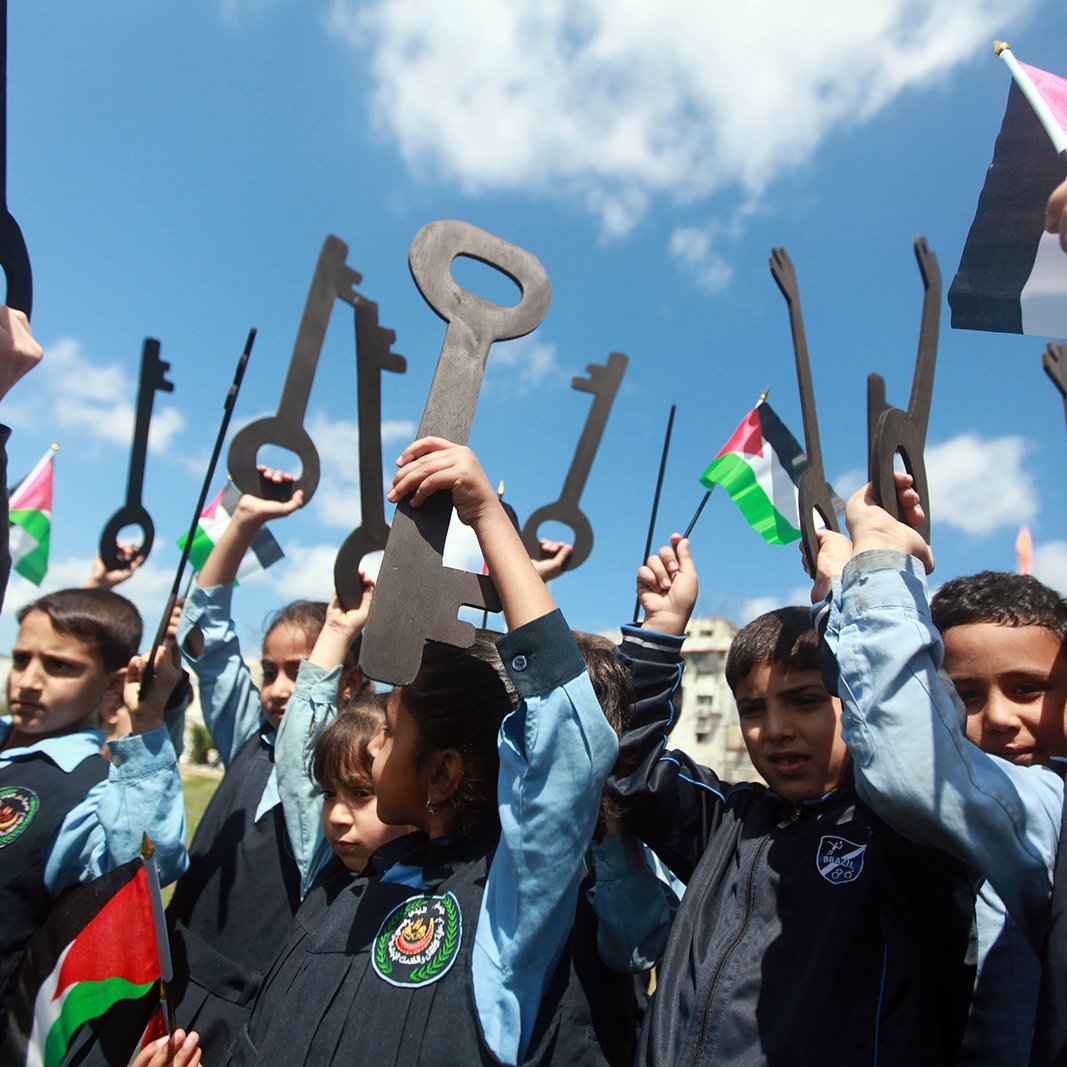 L'image montre un groupe d'enfants, qui semblent être en train de célébrer ou de manifester. Ils tiennent des grandes clés en carton et des drapeaux palestiniens. Les enfants, vêtus de uniformes scolaires, sourient et lèvent les clés vers le ciel, symbolisant un message de revendication ou d'espoir. Le ciel est bleu avec quelques nuages, ce qui ajoute à l'ambiance de la scène.