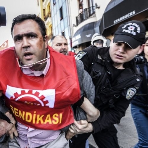 L'image montre une scène de répression où des policiers en tenue d'intervention arrêtent un homme qui porte un t-shirt rouge avec le mot "SENDIKASI". L'individu semble être en train d'être mené à l'écart, visiblement en désaccord avec son arrestation. Les policiers sont équipés de casques et semblent concentrés sur leur opération. L'environnement est urbain, et on peut voir un café Starbucks en arrière-plan, ce qui suggère que cette scène se déroule dans une zone animée de la ville.