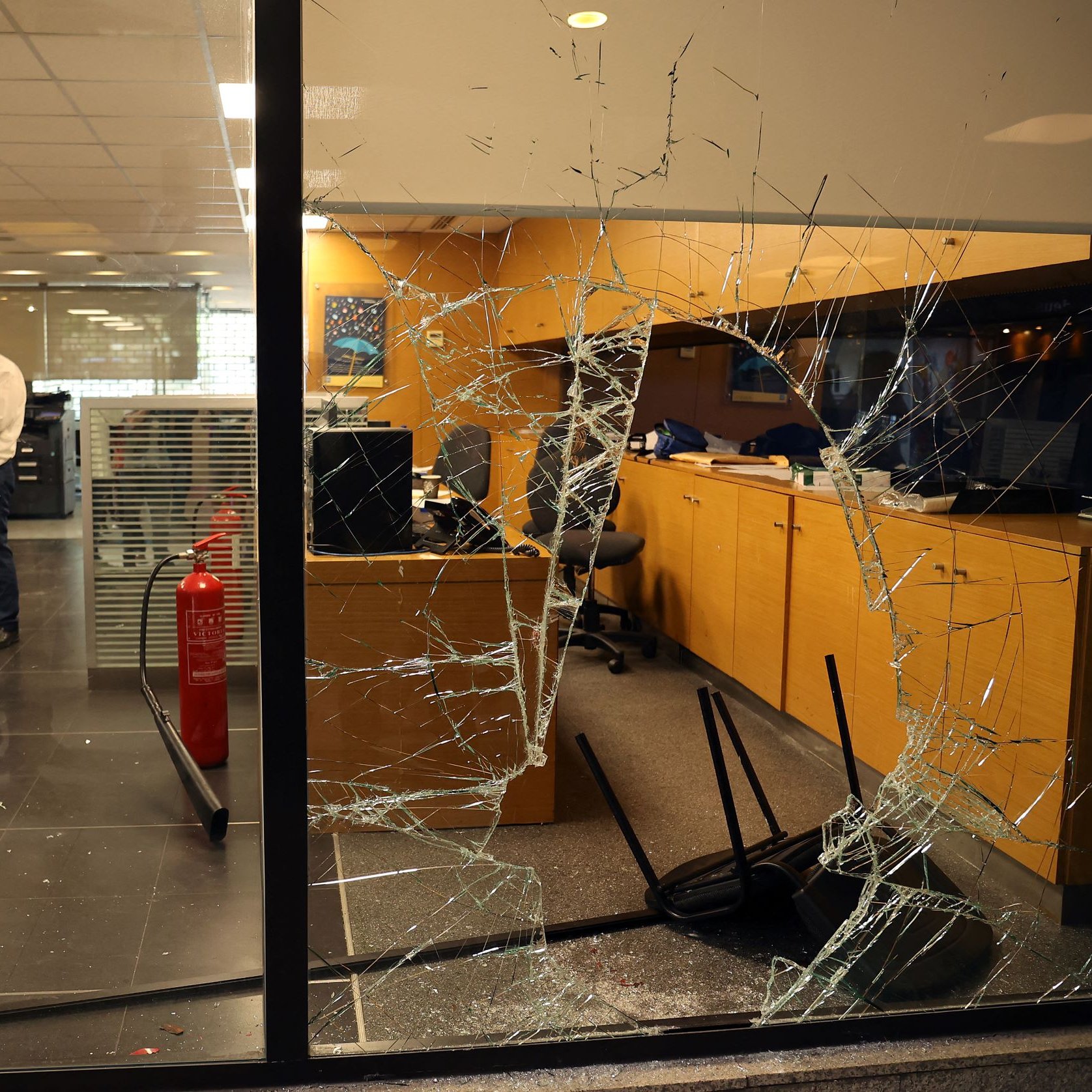 تظهر الصورة داخل مكتب يتضمن مشاهد من الفوضى، حيث يوجد زجاج مكسور على أحد النوافذ، مما يشير إلى وجود حادث أو تخريب. في الخلفية، يمكن رؤية أثاث مكتبي، مثل الطاولات والكراسي، وبعضها مقلوب. يوجد أيضًا شخص واحد يرتدي قميصًا فاتح اللون، يبدو وكأنه يتفقد الوضع داخل المكتب. الجو العام يوحي بحالة من الاضطراب أو الهجوم.