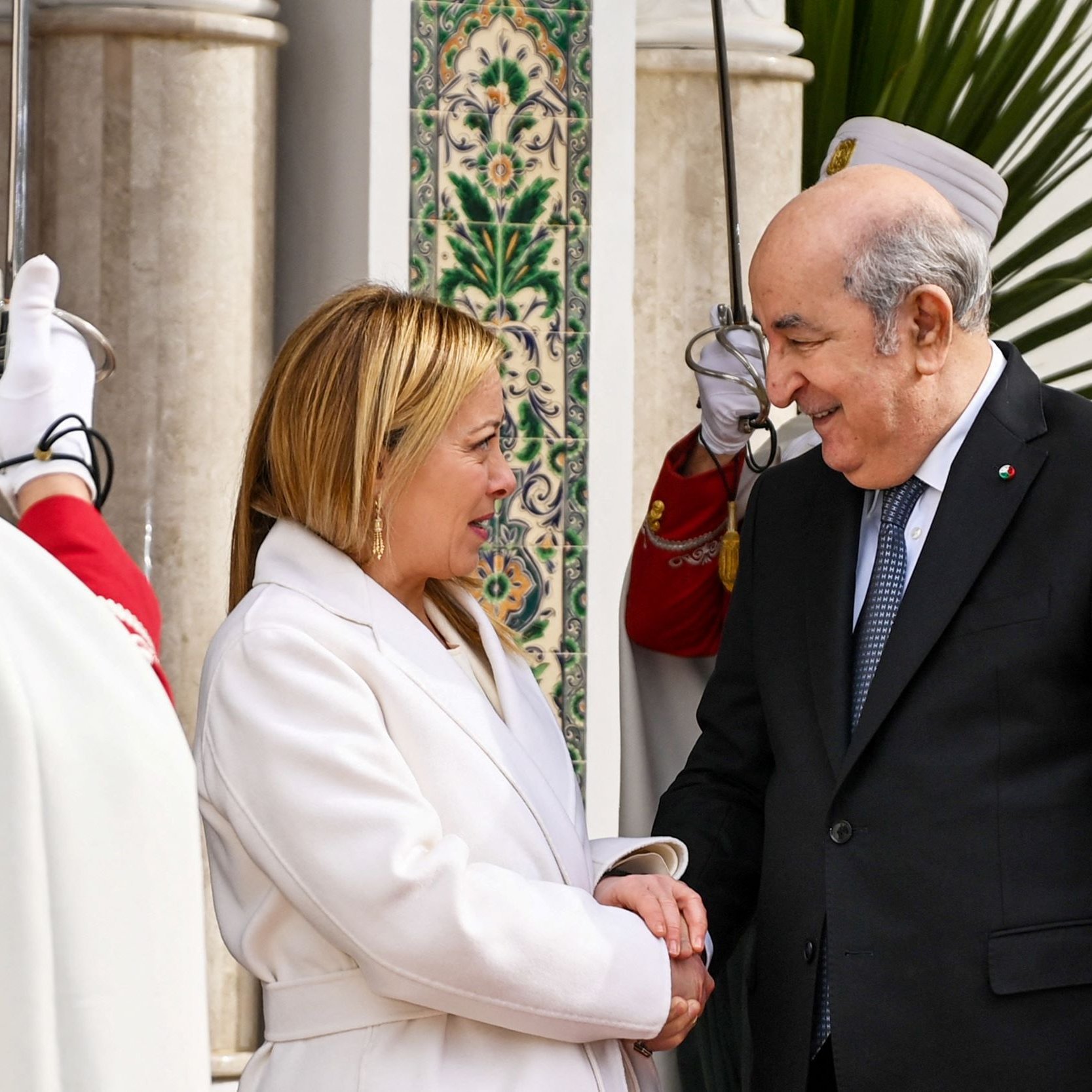 L'image montre deux personnes se serrant la main avec des sourires. Une femme, habillée en blanc, semble accueillante, tandis qu'un homme en costume sombre lui répond avec un sourire. En arrière-plan, on peut voir des gardes en uniforme et des éléments d'architecture décorative, ce qui suggère un contexte officiel, probablement une rencontre diplomatique. L'atmosphère est chaleureuse et conviviale.
