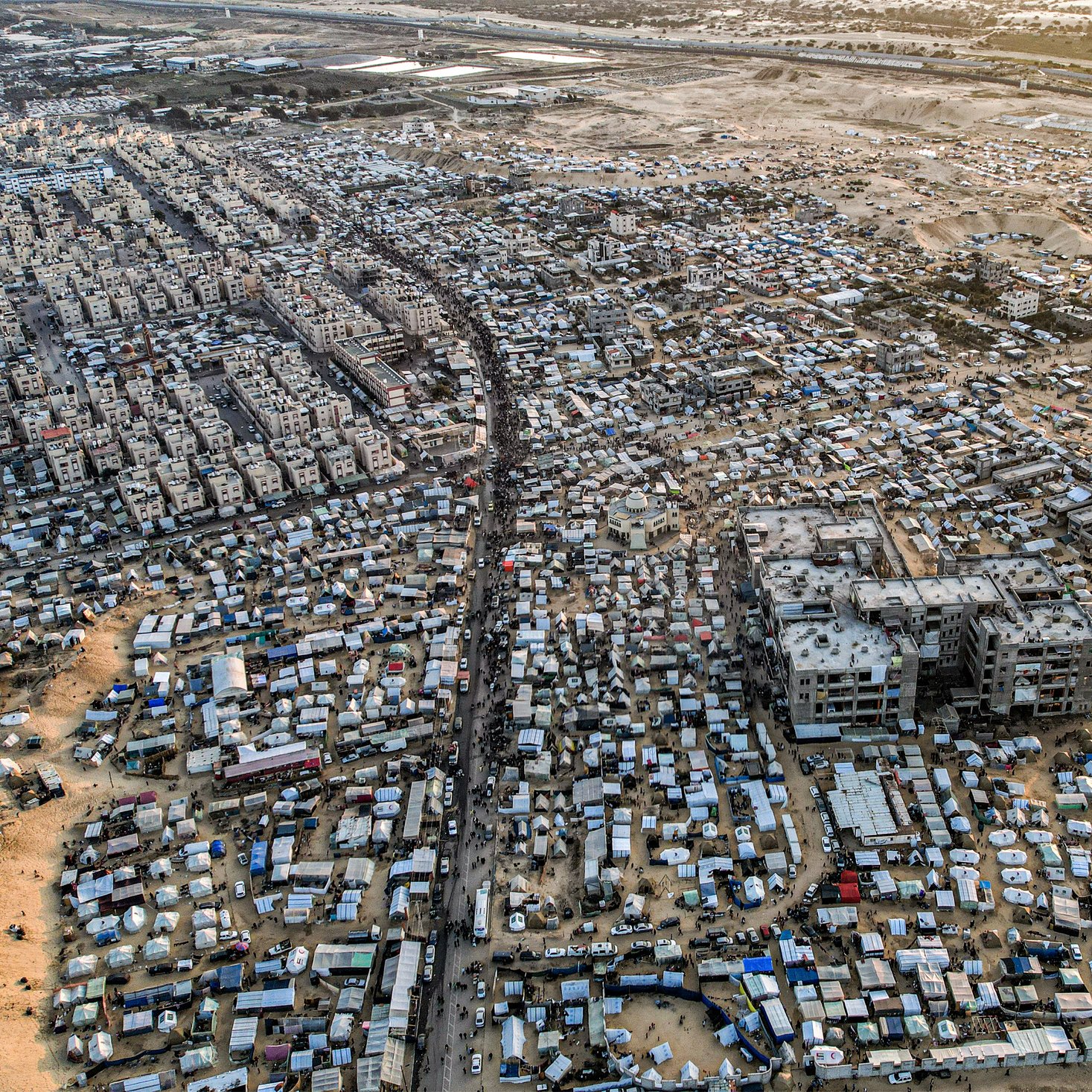 L'image montre une vue aérienne d'une vaste zone urbaine très densément peuplée, probablement une ville en développement ou un bidonville. On peut distinguer des habitations de différentes tailles et styles, de nombreuses structures temporaires, ainsi que des routes et des chemins qui traversent l'ensemble de la zone. La scène est marquée par un mélange de constructions durables et de tentes, suggérant une forte densité de population ainsi que des conditions de vie variées. En arrière-plan, on aperçoit des terrains plus dégagés, peut-être des zones agricoles ou industrielles.