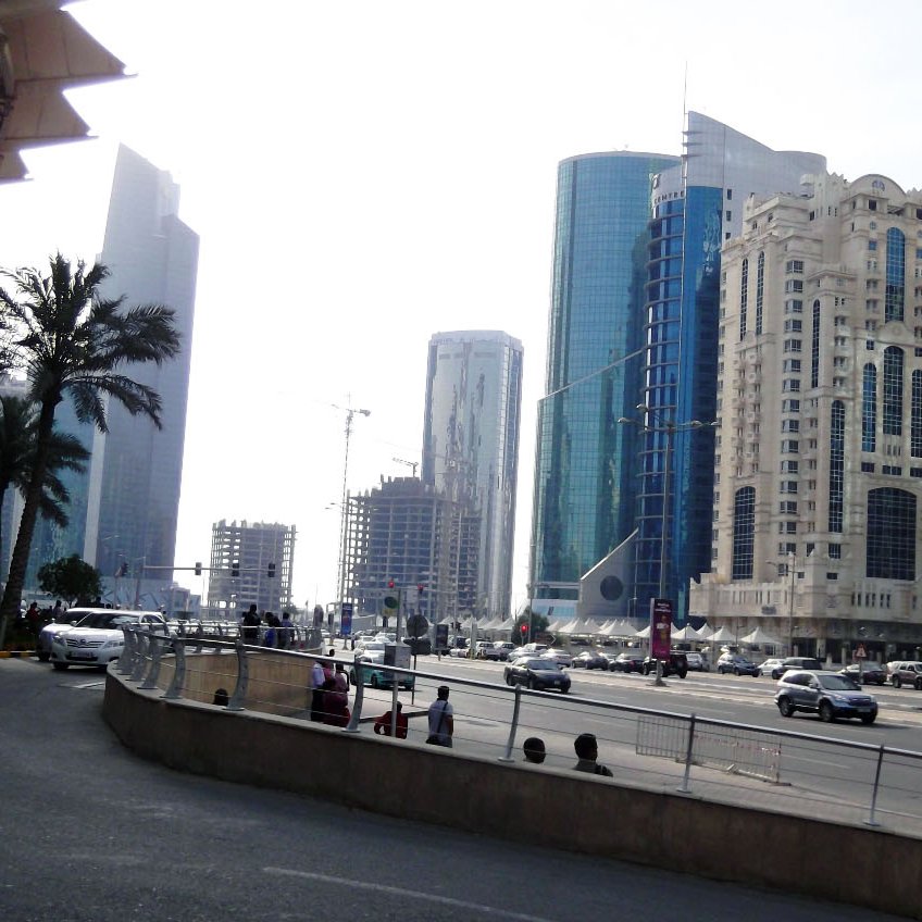 The image depicts a busy city street featuring a mix of modern skyscrapers and architectural styles. On the left, there are palm trees and a wide road leading into the city. Various cars are seen driving, indicating active traffic. The tall buildings on either side showcase glass and steel facades, reflecting light from the environment. The overall atmosphere suggests a bustling urban area with a contemporary vibe. The sky appears bright, contributing to the lively scene.