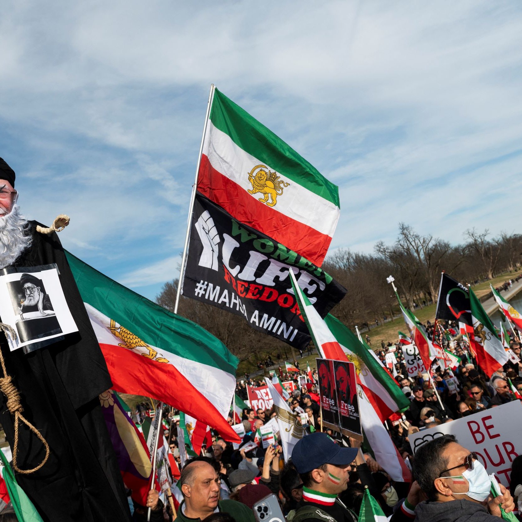 L'image montre une grande manifestation, probablement en faveur de la liberté ou des droits de l'homme. Au premier plan, une marionnette représentant un homme âgé est portée par un manifestant, tandis que d'autres personnes tiennent des drapeaux iraniens et des pancartes. Les participants semblent unis et déterminés, exprimant leurs revendications. L'ambiance est dynamique, avec une foule nombreuse et des symboles visuels forts liés à leur cause.