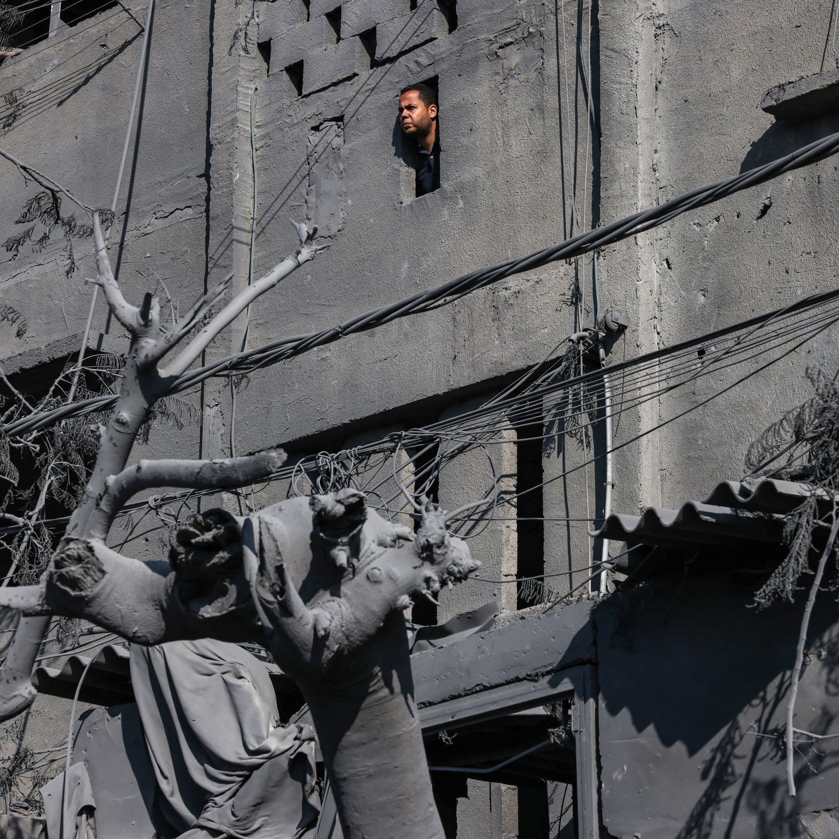 L'immagine mostra un edificio gravemente danneggiato, con i muri di cemento visibilmente rovinati. Da una finestra rotta, si intravede una persona che osserva l'esterno, mentre nel primo piano si trova una statua, anch'essa coperta di polvere e detriti. Le linee elettriche pendono disordinatamente, e l'atmosfera è cupa a causa dei colori grigi e della desolazione del contesto. La scena sembra suggerire un ambiente colpito da un evento catastrofico, con evidenti segni di distruzione e abbandono.