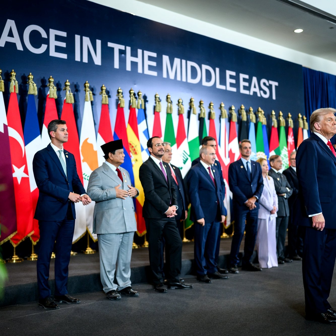 A group of leaders stands in front of flags, with one man speaking, promoting Middle East peace.
