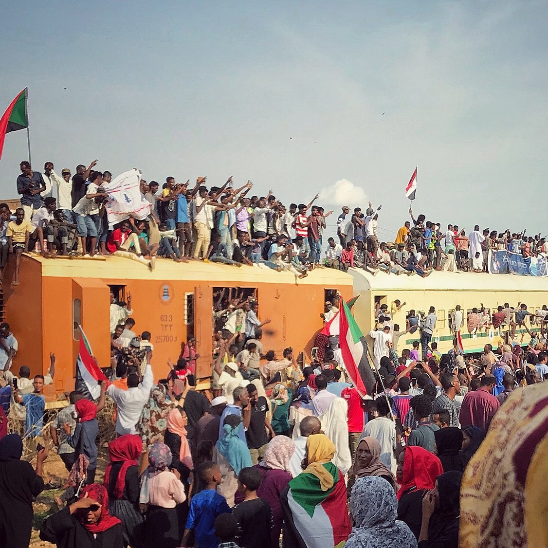 L'image montre une grande foule de manifestants rassemblés autour d'un train. De nombreuses personnes sont sur le toit et les côtés du train, brandissant des drapeaux, notamment des drapeaux soudanais. L'atmosphère semble énergique et festive, avec des gens qui expriment leur enthousiasme et leur solidarité. Le ciel est partiellement nuageux, et la scène donne une impression de mouvement et d'engagement collectif.