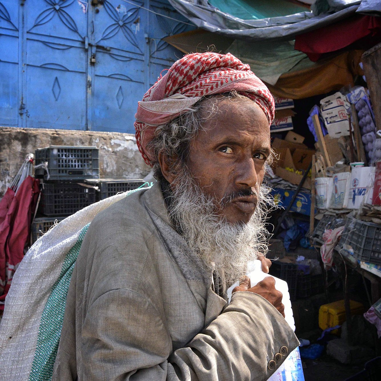 L'image montre un homme âgé avec une longue barbe grise, portant un turban rouge et blanc. Il semble regarder fixement avec une expression pensante ou préoccupée. L'arrière-plan suggère un marché ou un bazar, avec des étals et des objets divers. L'éclairage est naturel, ce qui donne une atmosphère vivante à la scène. L'homme porte un sac, ce qui indique qu'il pourrait être en train de se déplacer ou de faire des courses.