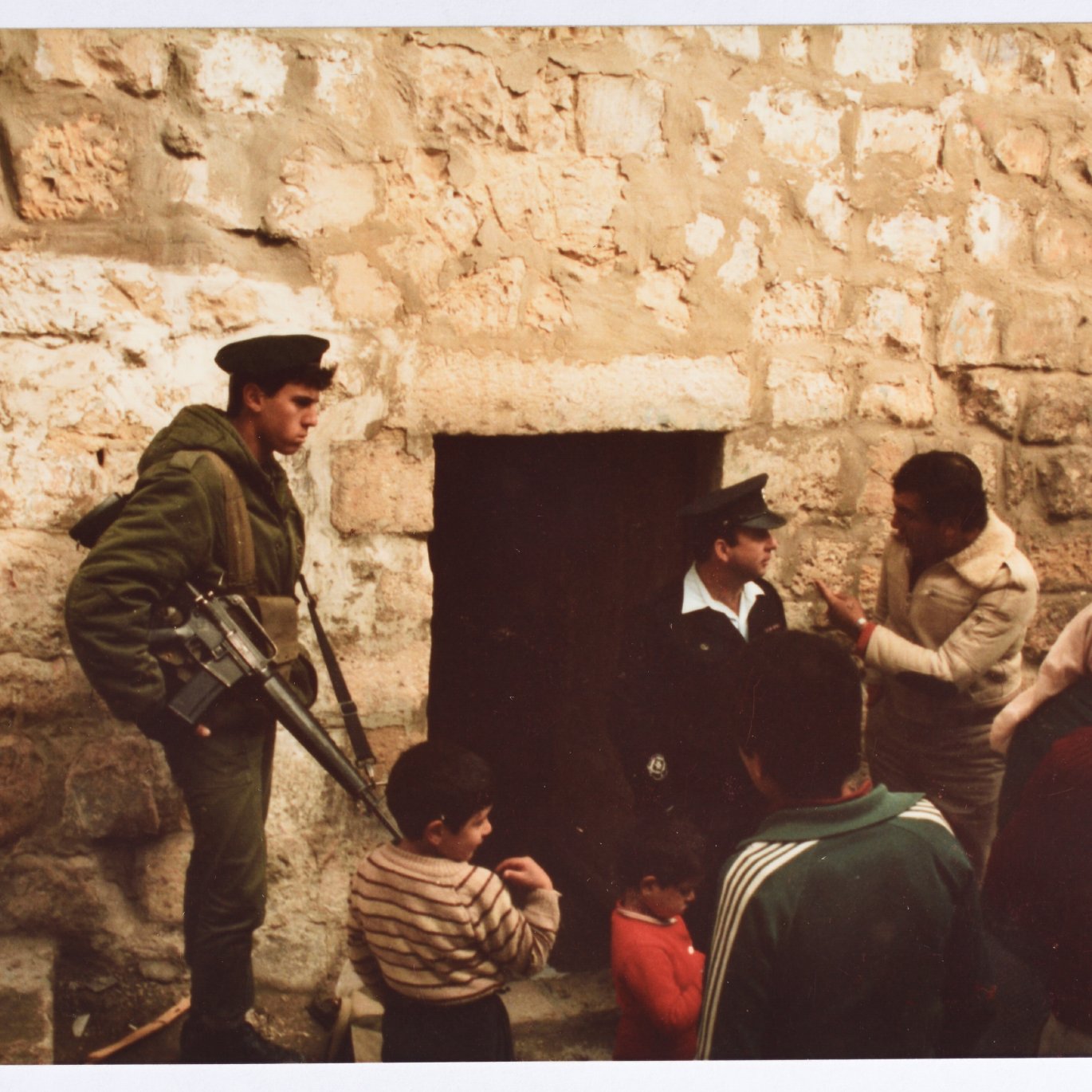 L'image montre un groupe de personnes près d'une porte en pierre. À gauche, un soldat armé en uniforme vert se tient debout, gardant l'entrée. Deux hommes adultes discutent, tandis qu'un enfant et un autre homme semblent observer la scène. L'environnement est marqué par des murs de pierre brute, suggérant un endroit ancien ou historique. L'atmosphère paraît chargée, et les expressions des personnes indiquent une interaction sérieuse.