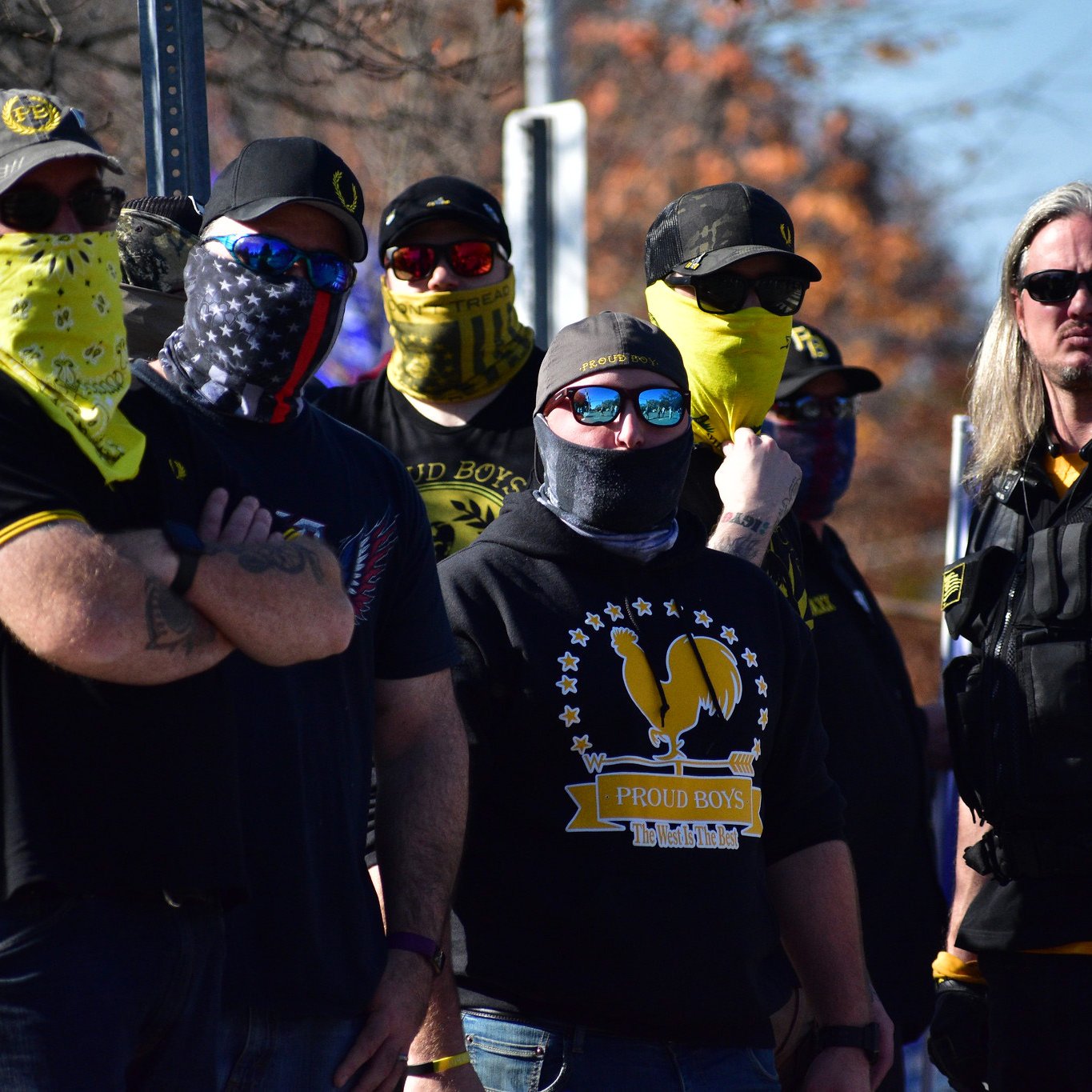 L'image montre un groupe de personnes portant des vêtements sombres, accompagnés de foulards ou de masques qui cachent en partie leur visage. Ils se tiennent en ligne, affichant des expressions sérieuses et une posture déterminée. Certains portent des lunettes de soleil et ont des logos visibles sur leurs vêtements. L'environnement semble être en extérieur, avec des arbres et un panneau visible en arrière-plan. L'atmosphère semble tendue, avec une présence marquée de camaraderie parmi le groupe.