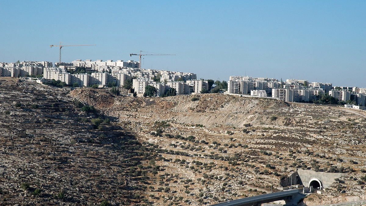 L'image montre un paysage caractérisé par une colline aride et rocheuse, sur laquelle se trouvent des bâtiments modernes. En arrière-plan, on peut voir des constructions en cours, avec des grues qui ajoutent à l'atmosphère de développement urbain. Au premier plan, une structure en béton est visible, semblant faire partie d'une voie ou d'un pont qui traverse le terrain. Le ciel est dégagé, ce qui donne une impression de luminosité et d'espace.