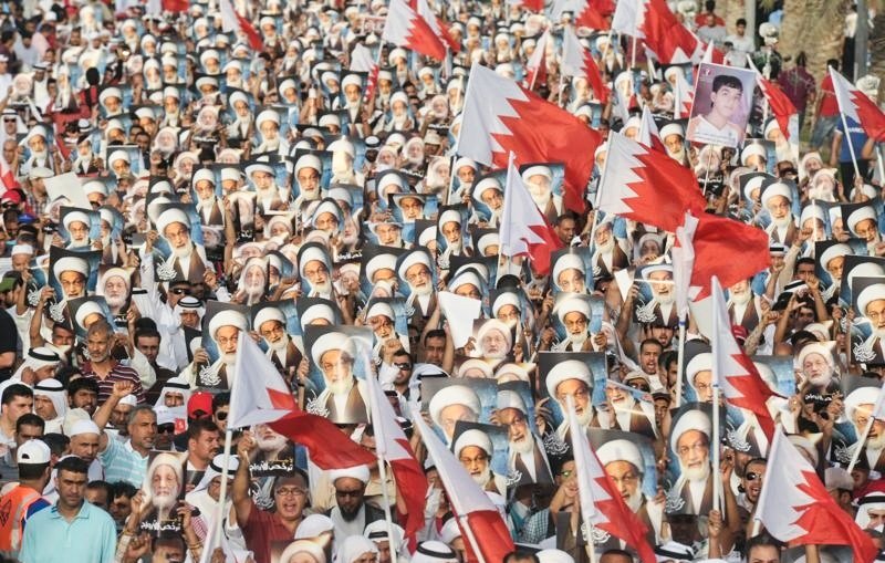 الصورة تُظهر تجمعاً حاشداً لمتظاهرين في البحرين. المشاركون يحملون أعلام البحرين، بالإضافة إلى صور لشخصيات معروفة. الجو يبدو حماسياً، حيث يرتدي العديد من المشاركين الثياب التقليدية. المناظر تعكس روح التضامن والمشاركة الفاعلة في الحدث.
