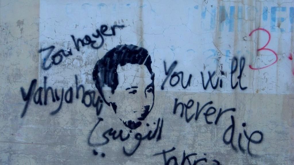 تظهر الصورة رسمًا على جدار يظهر وجه شخص، محاطًا بعبارات مكتوبة بخط يدوي. من بين هذه العبارات، تُظهر باللغة الإنجليزية "You will never die" (لن تموت أبدًا) وفي اللغة العربية "يحيى" و"تقديس". يبدو أن العمل يعبر عن رسالة تعاطف أو تأبين، مع تداخل بين الثقافات واللغات في الكتابة.